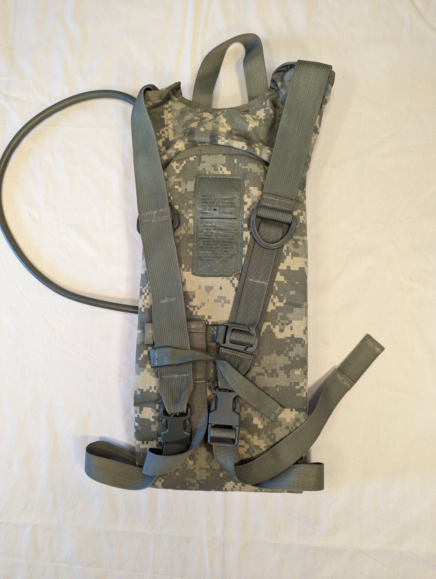 A front view of a U.S. Military Surplus Hydration Carrier in ACU digital camouflage, standing upright, featuring the attached drinking tube, shoulder straps, and quick-release buckles.