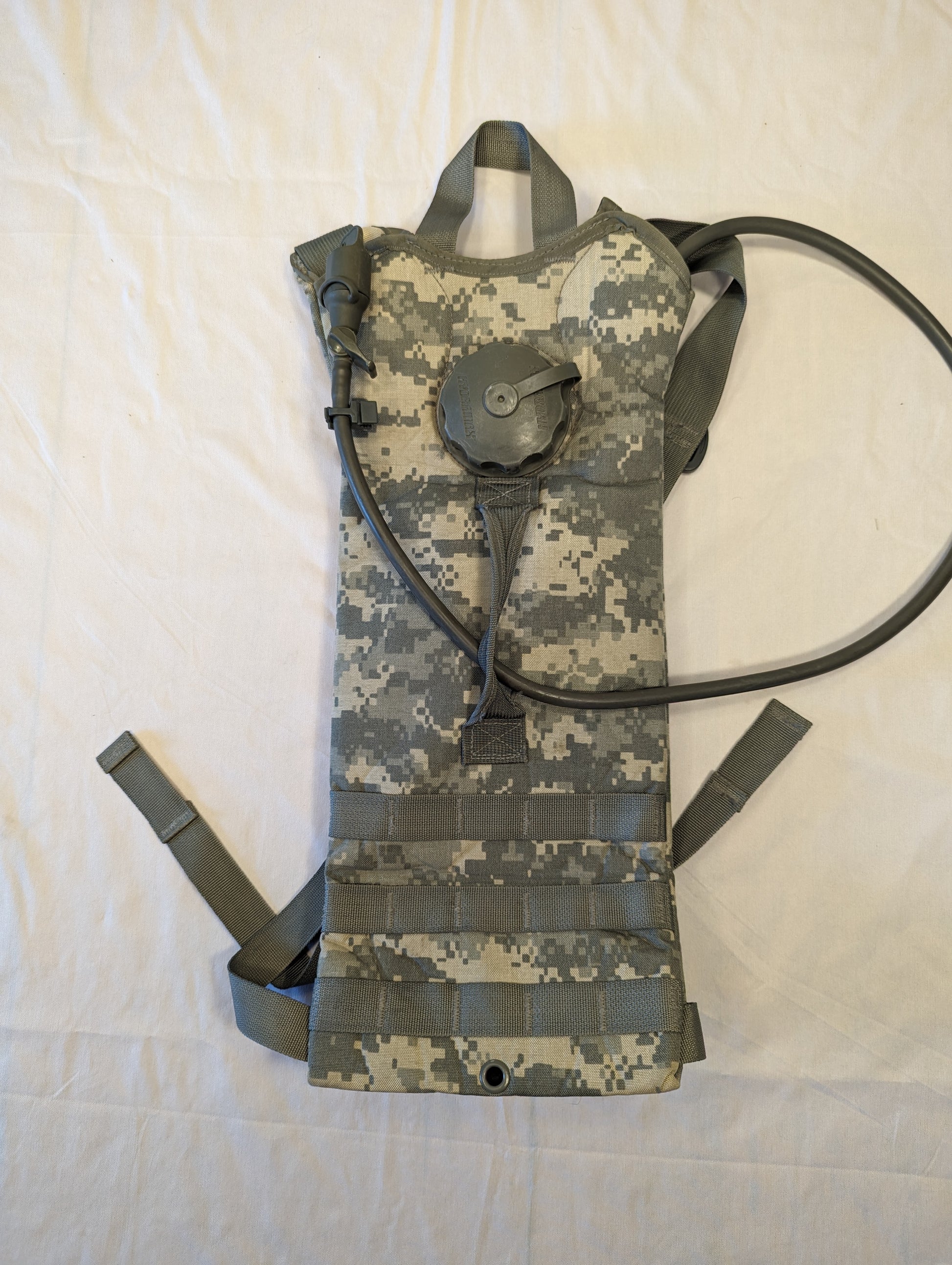 A front view of a U.S. Military Surplus Hydration Carrier in ACU digital camouflage, laid flat, featuring the MOLLE webbing straps, a carrying handle, and the attached drinking tube and cap.