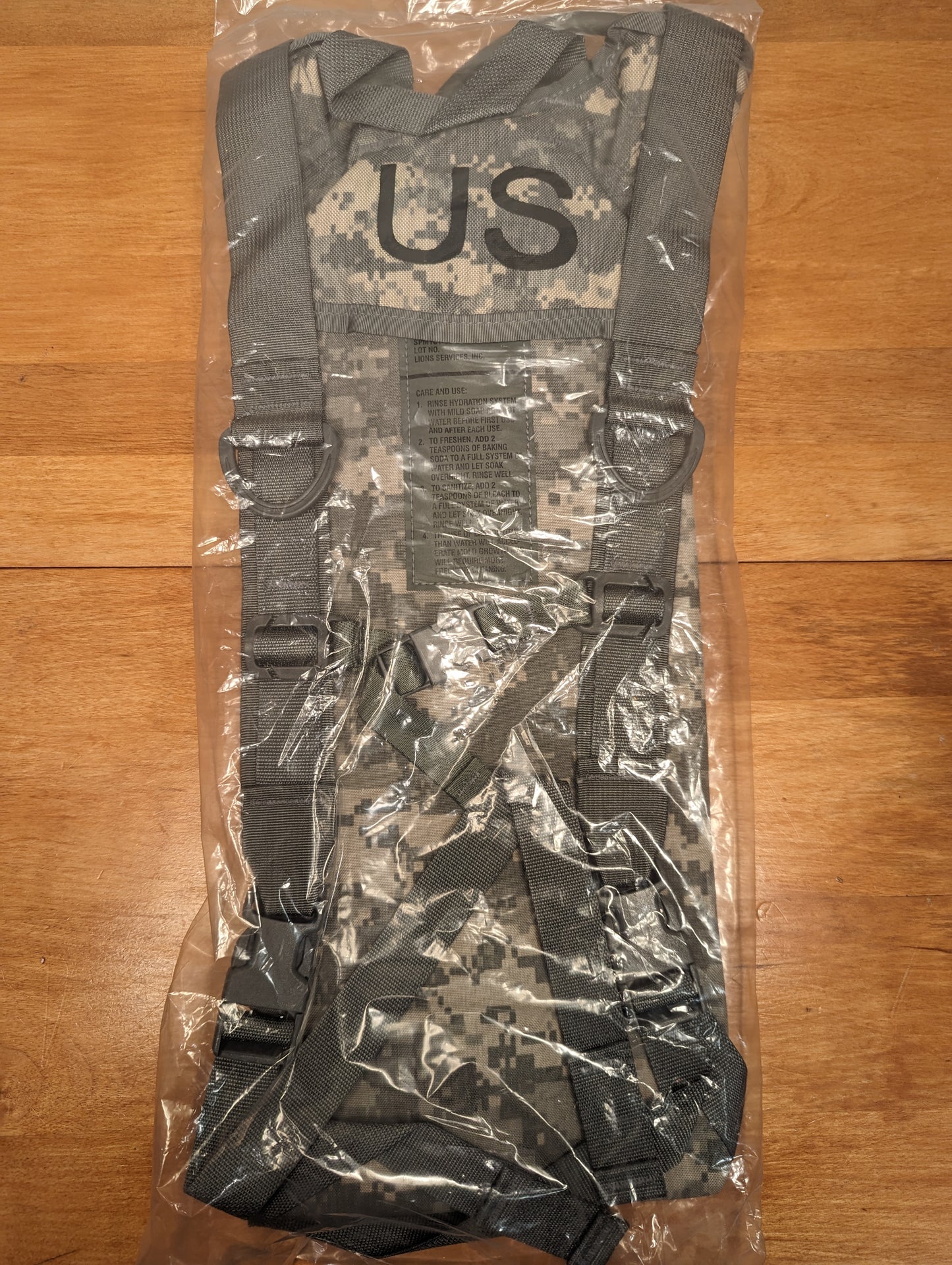 A new U.S. Military Surplus Hydration Carrier in ACU digital camouflage, sealed in clear plastic packaging and laid on a wood background, with the large 'US' military mark visible on the front.