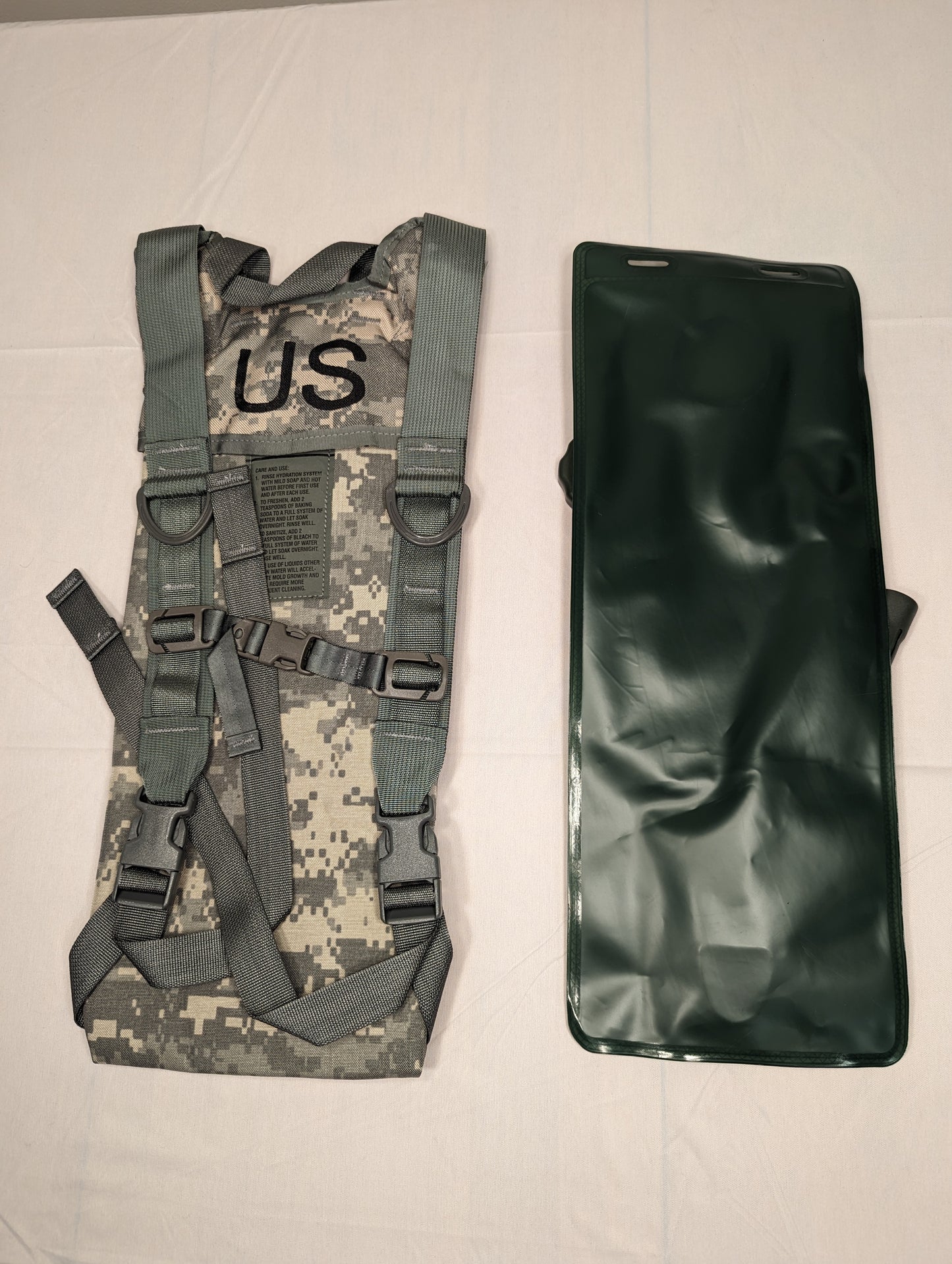 A studio shot of the U.S. Military Hydration Carrier components laid side-by-side: the ACU digital camouflage carrier shell with a stenciled 'US' mark, and the dark olive green hydration bladder.