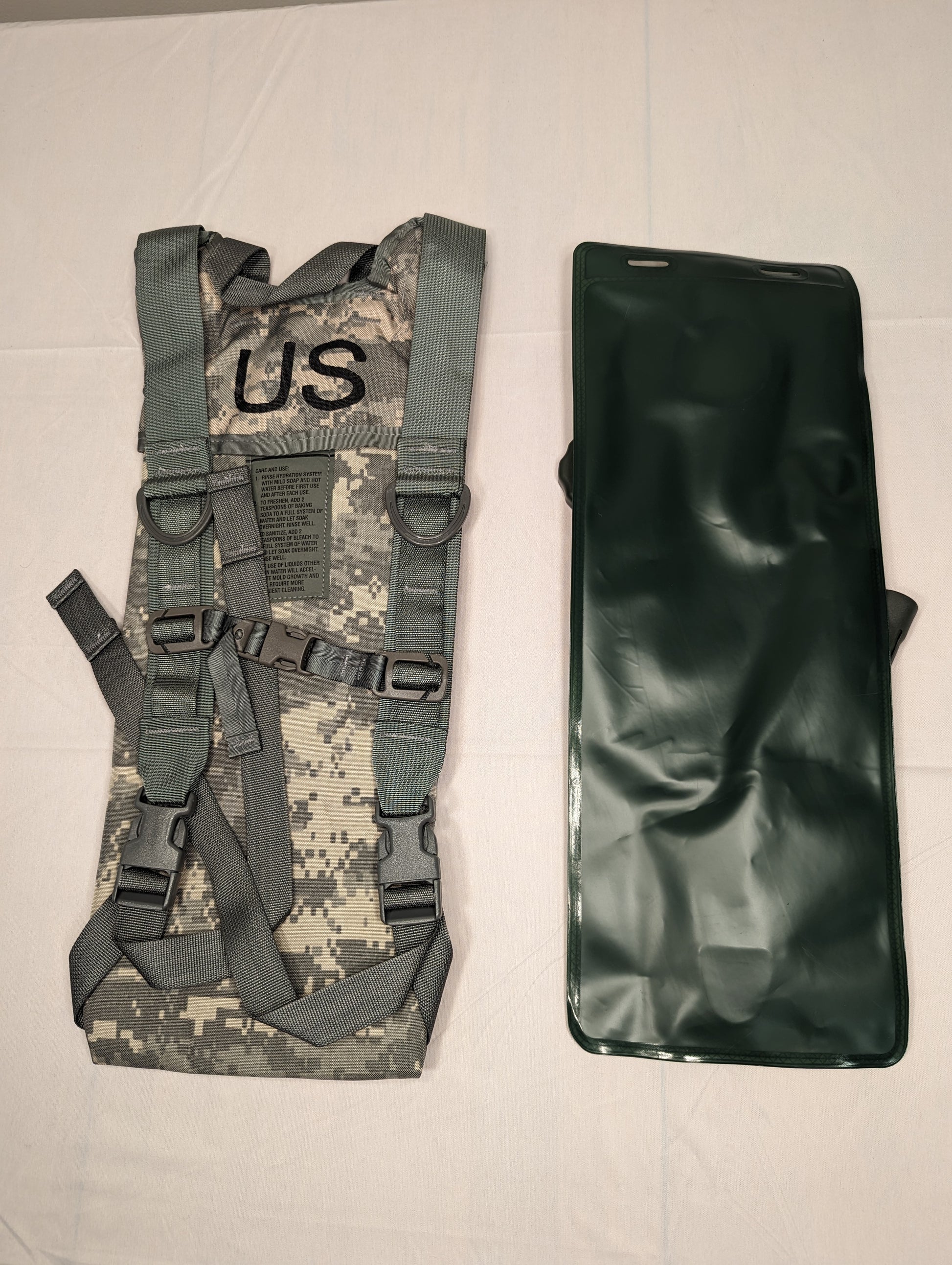 A studio shot of the U.S. Military Hydration Carrier components laid side-by-side: the ACU digital camouflage carrier shell with a stenciled 'US' mark, and the dark olive green hydration bladder.