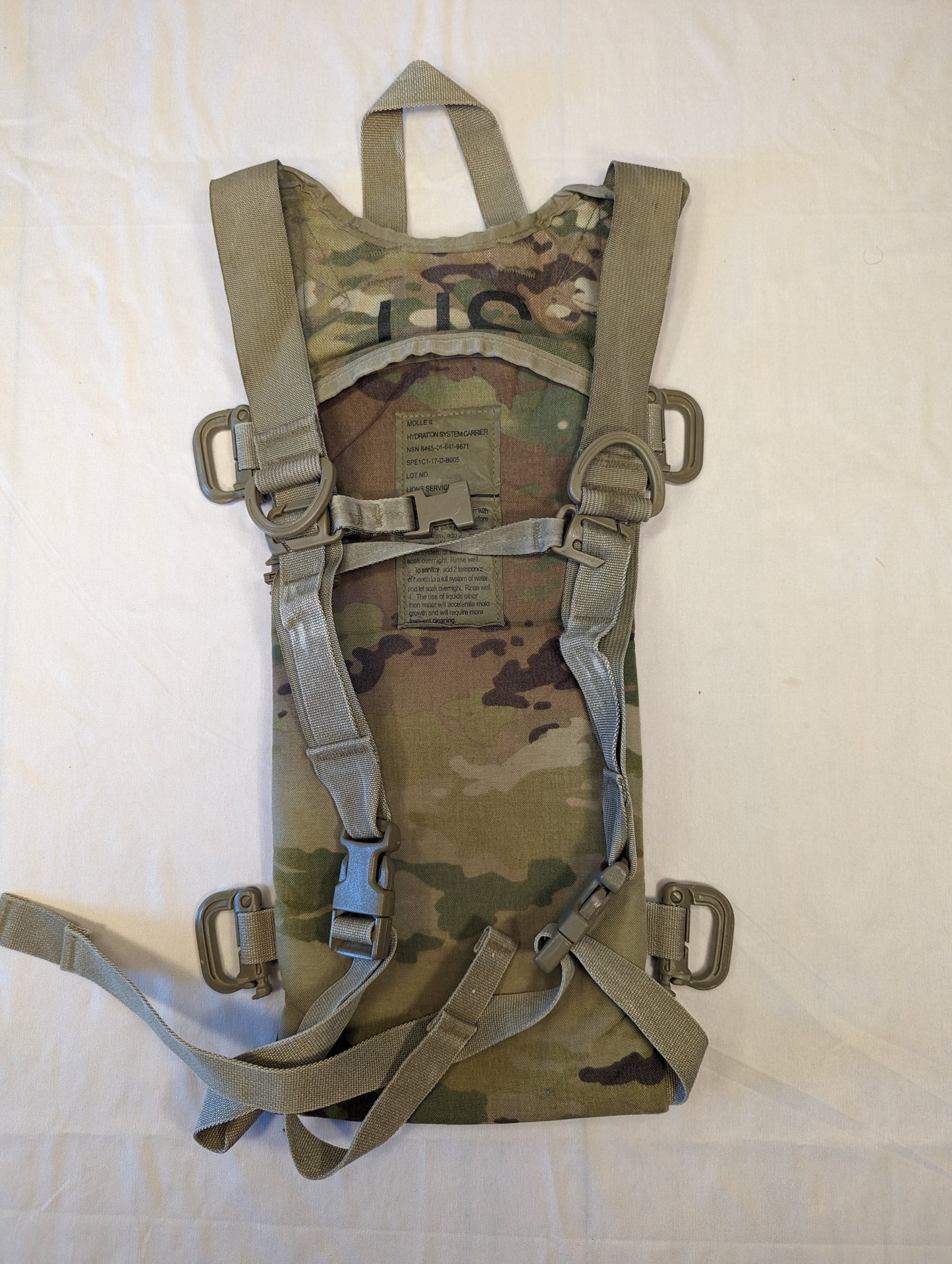 A back view of a U.S. Military Surplus Hydration Carrier in OCP/Multicam camouflage, laid flat, showcasing the full padded harness system, tan quick-release buckles, D-rings, and visible military issue tag.