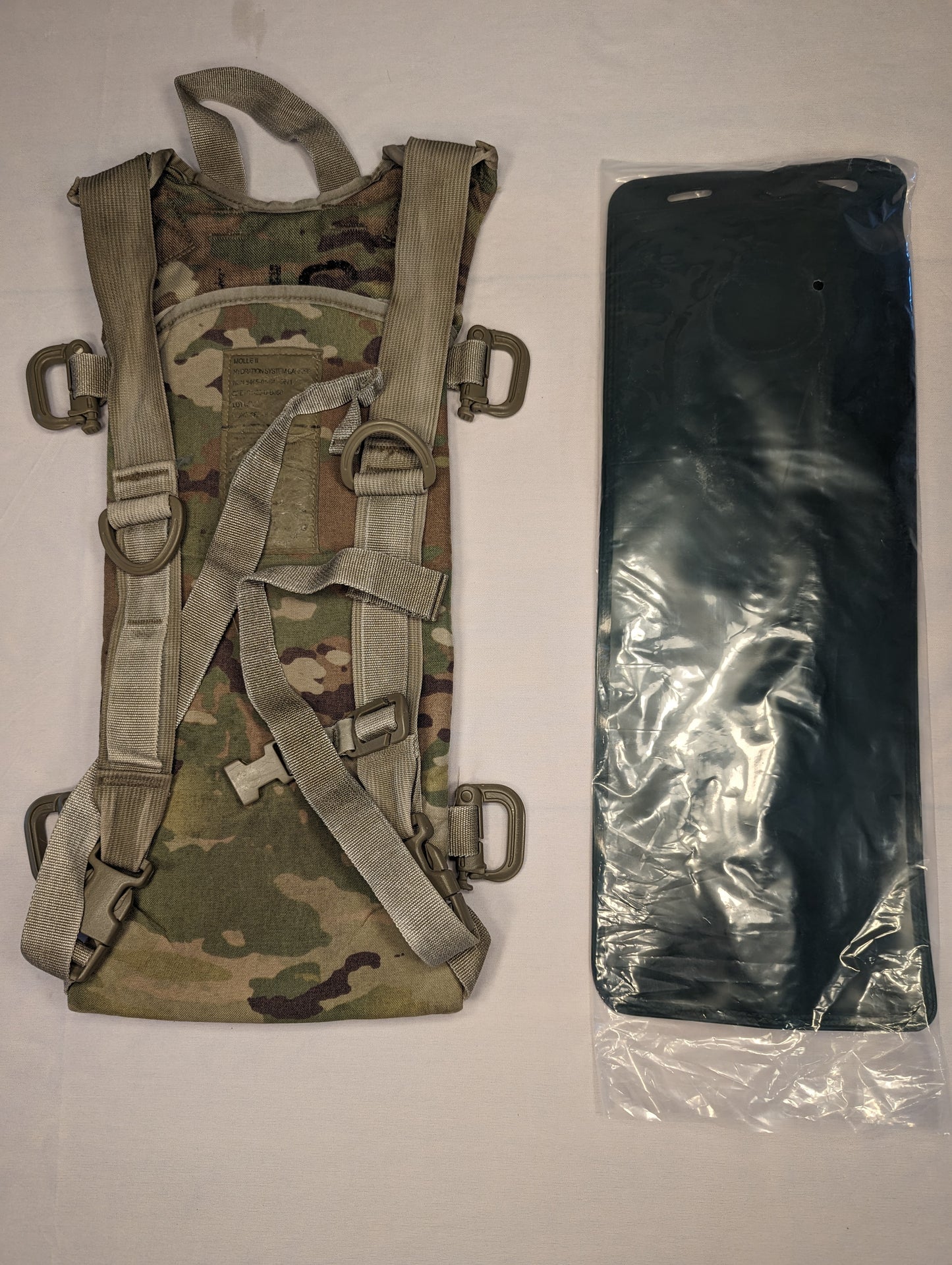 A U.S. Military Surplus Hydration Carrier in OCP/Multicam camouflage, laid flat, with its MOLLE webbing visible, displayed next to a new, black, sealed hydration bladder.