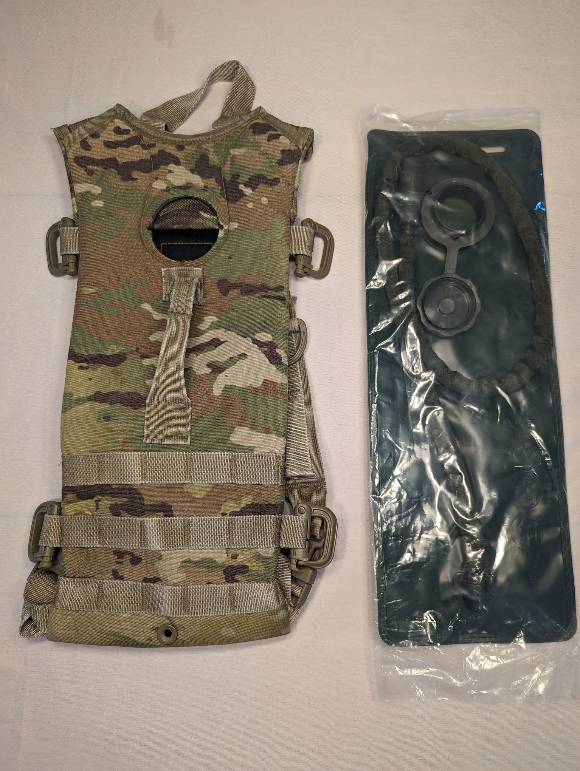 A U.S. Military Surplus Hydration Carrier in OCP/Multicam camouflage, laid flat, with its MOLLE webbing visible, displayed next to a new, black, sealed hydration bladder.