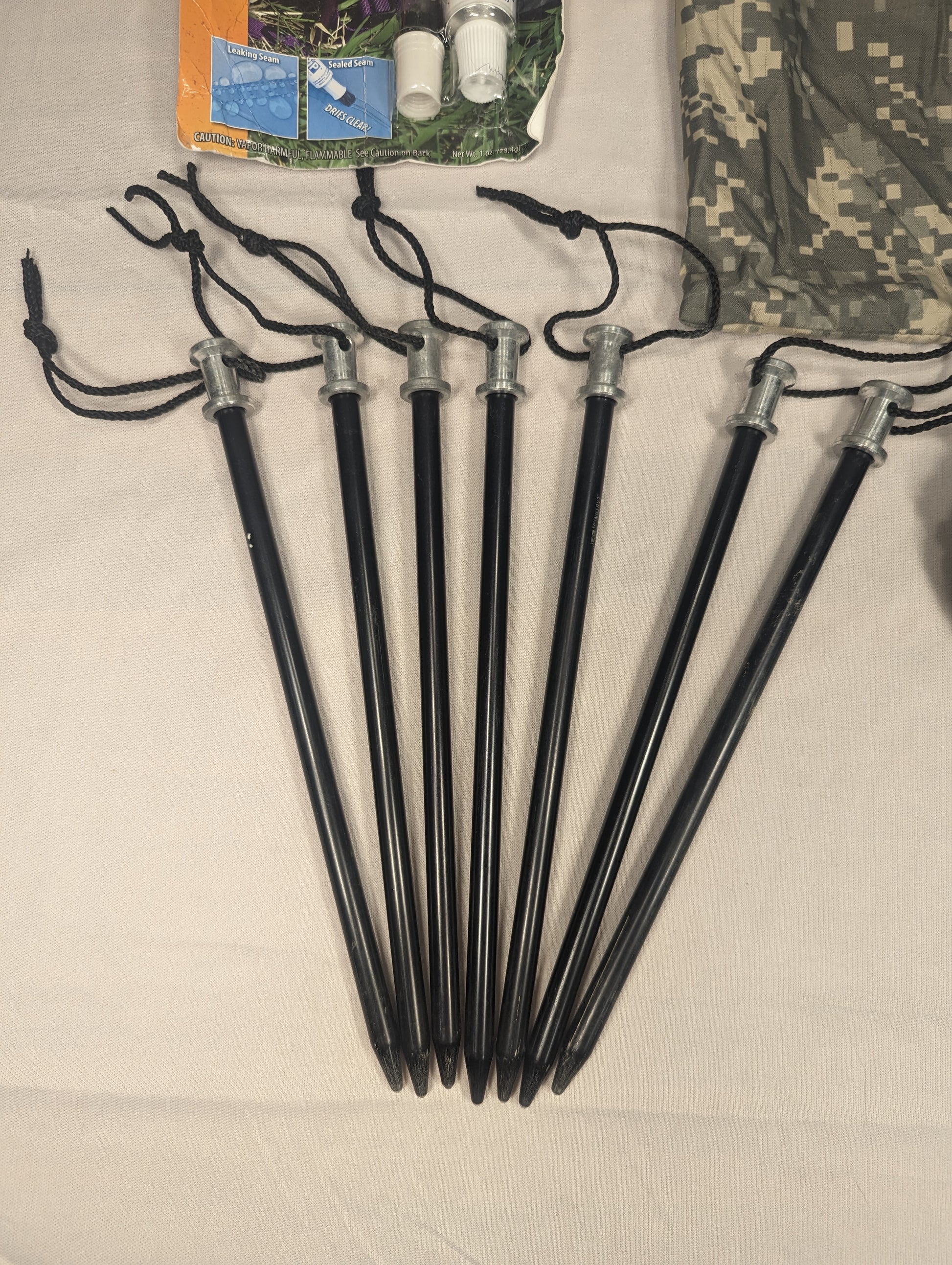 A set of seven dark-colored tent stakes with black paracord loops, arranged in a fan shape, part of the ICS Repair Kit, with camouflage fabric and sealant visible in the background.