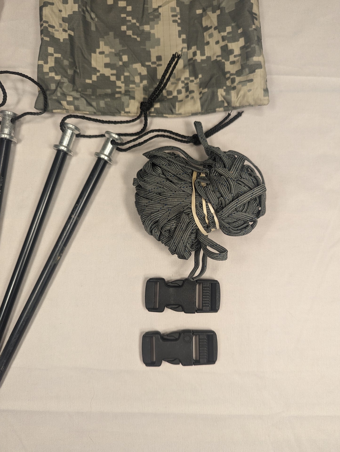 A close-up of a U.S. Military ICS Tent Repair Kit components, featuring three black tent poles with paracord, a coil of gray rope, and two black quick-release buckles, laid next to ACU digital camouflage fabric.