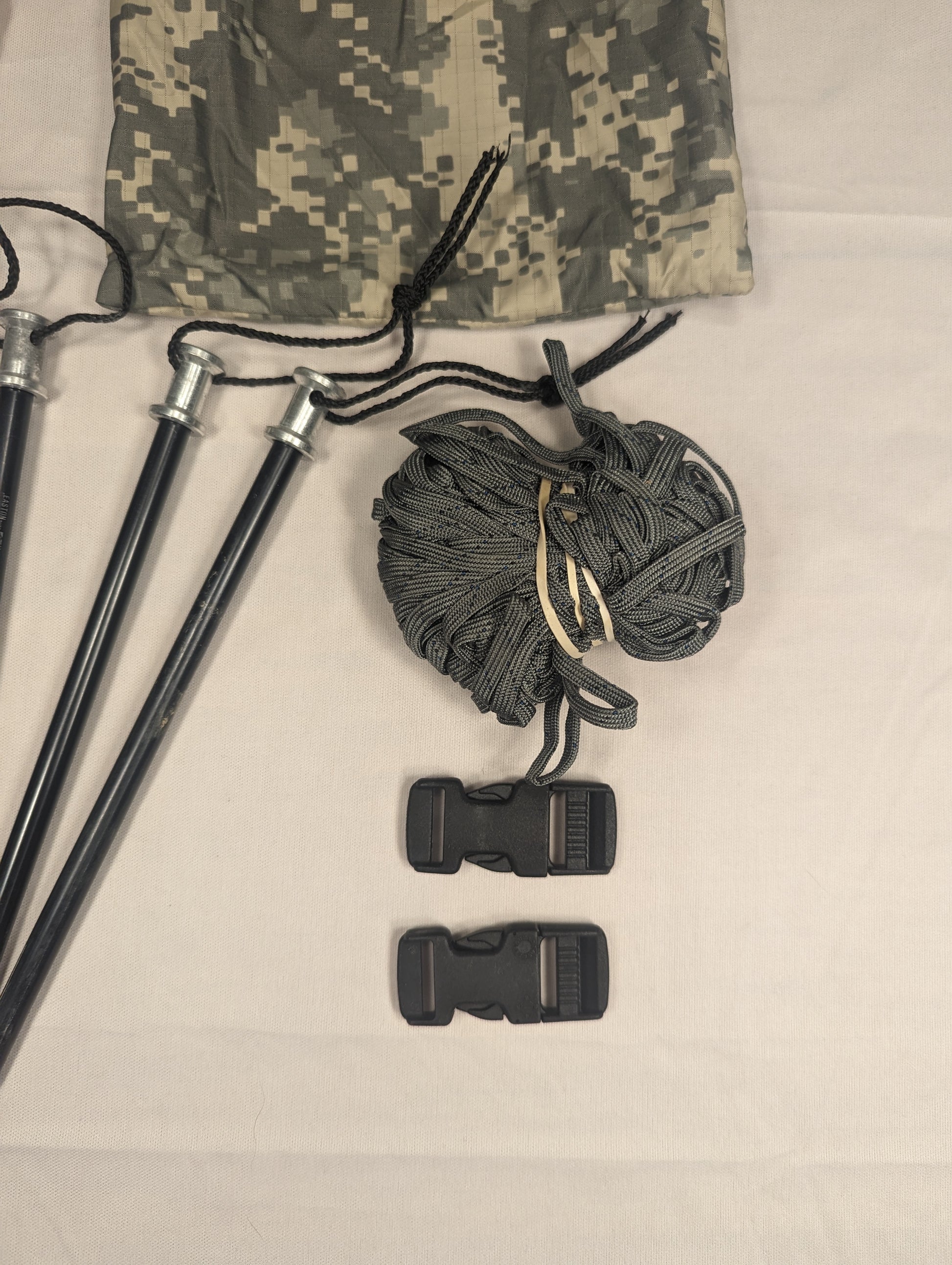 A close-up of a U.S. Military ICS Tent Repair Kit components, featuring three black tent poles with paracord, a coil of gray rope, and two black quick-release buckles, laid next to ACU digital camouflage fabric.