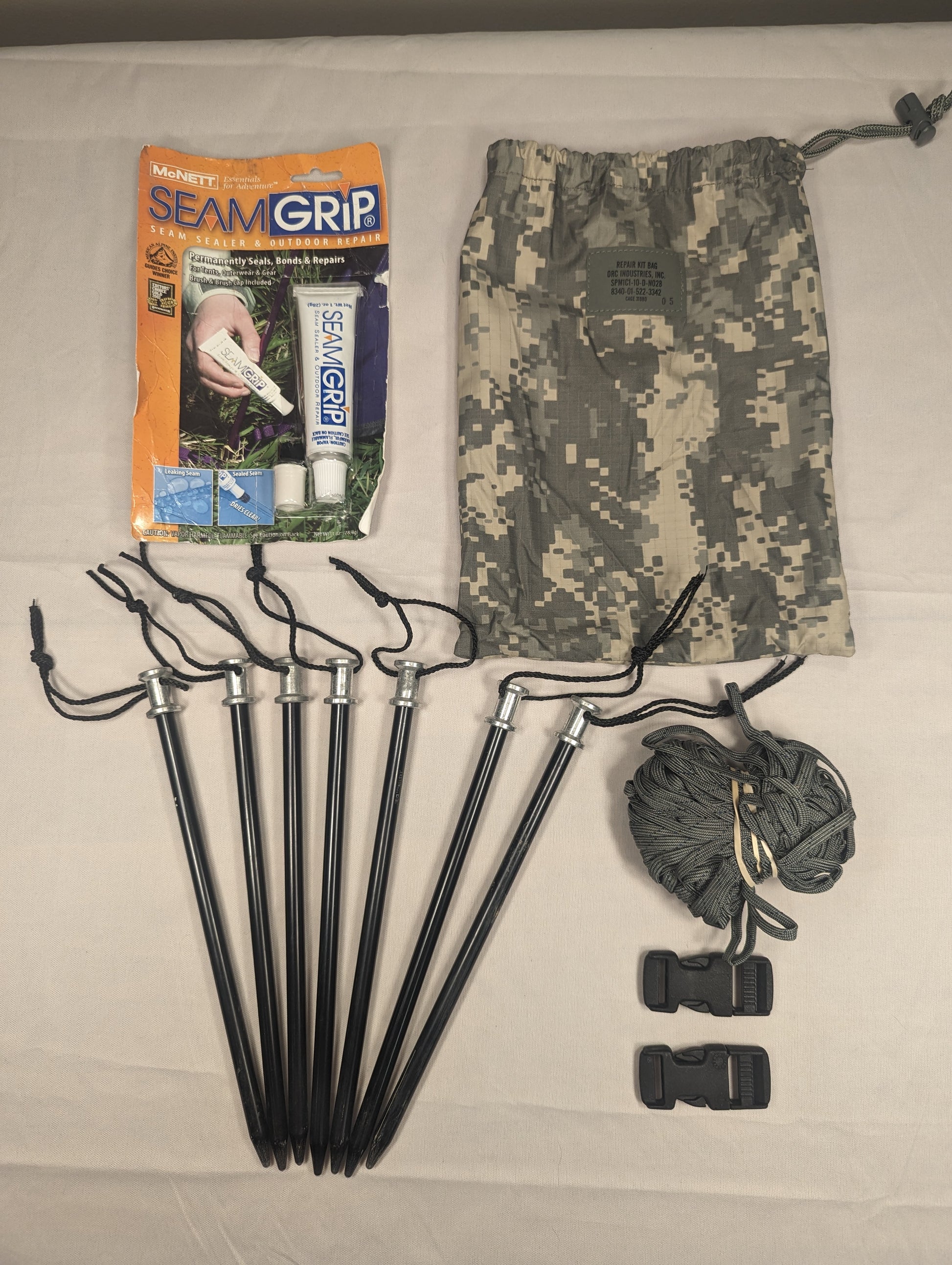 An overhead view of a U.S. Military tent repair kit laid out flat, including seven dark tent stakes with cord, a camouflage drawstring pouch, a tube of repair sealant, and two plastic quick-release buckles.