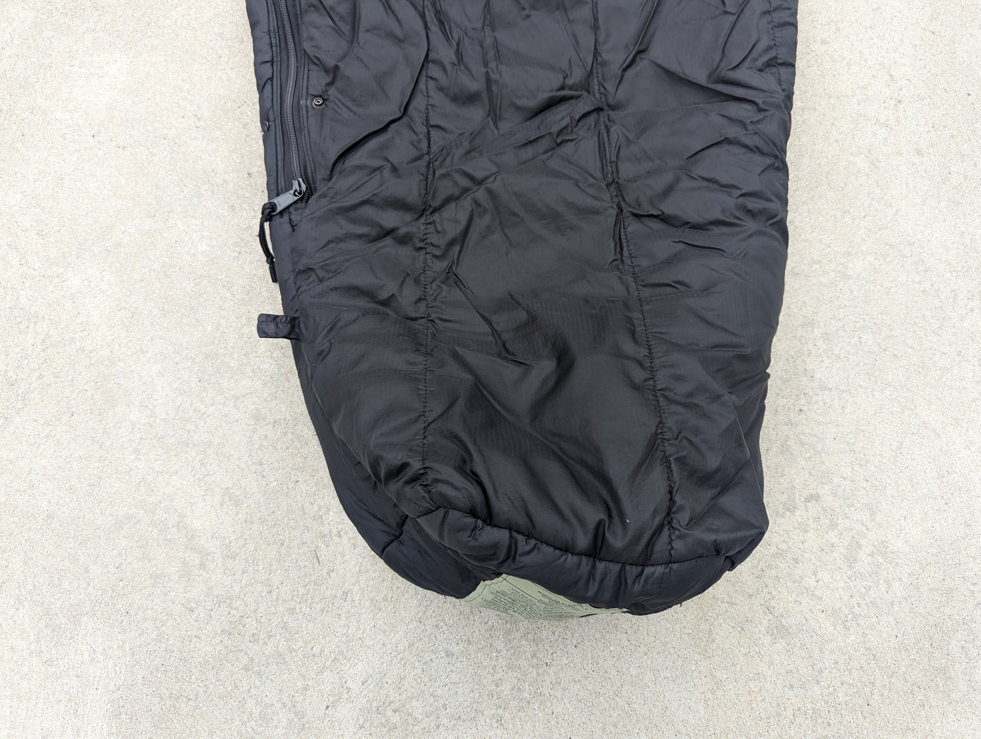A close-up, top-down view of the foot box of a U.S. Military Intermediate Cold Weather Sleeping Bag in black, showing the quilted nylon fabric, zipper closure, and olive green bottom section.