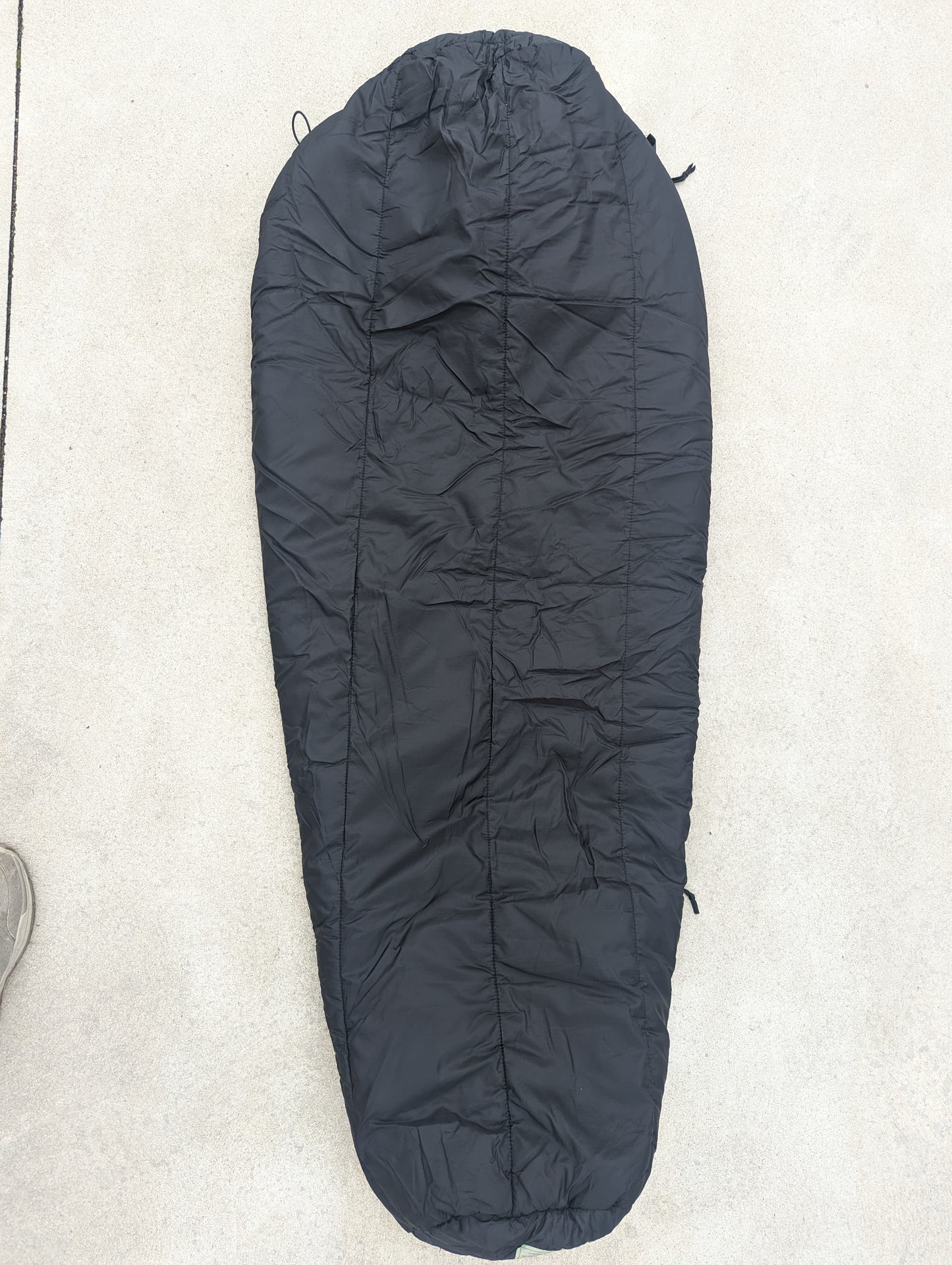 A full-length U.S. Military Surplus Intermediate Cold Weather Sleeping Bag in black, zipped and cinched closed, displaying its bulky, quilted mummy shape on a light background.