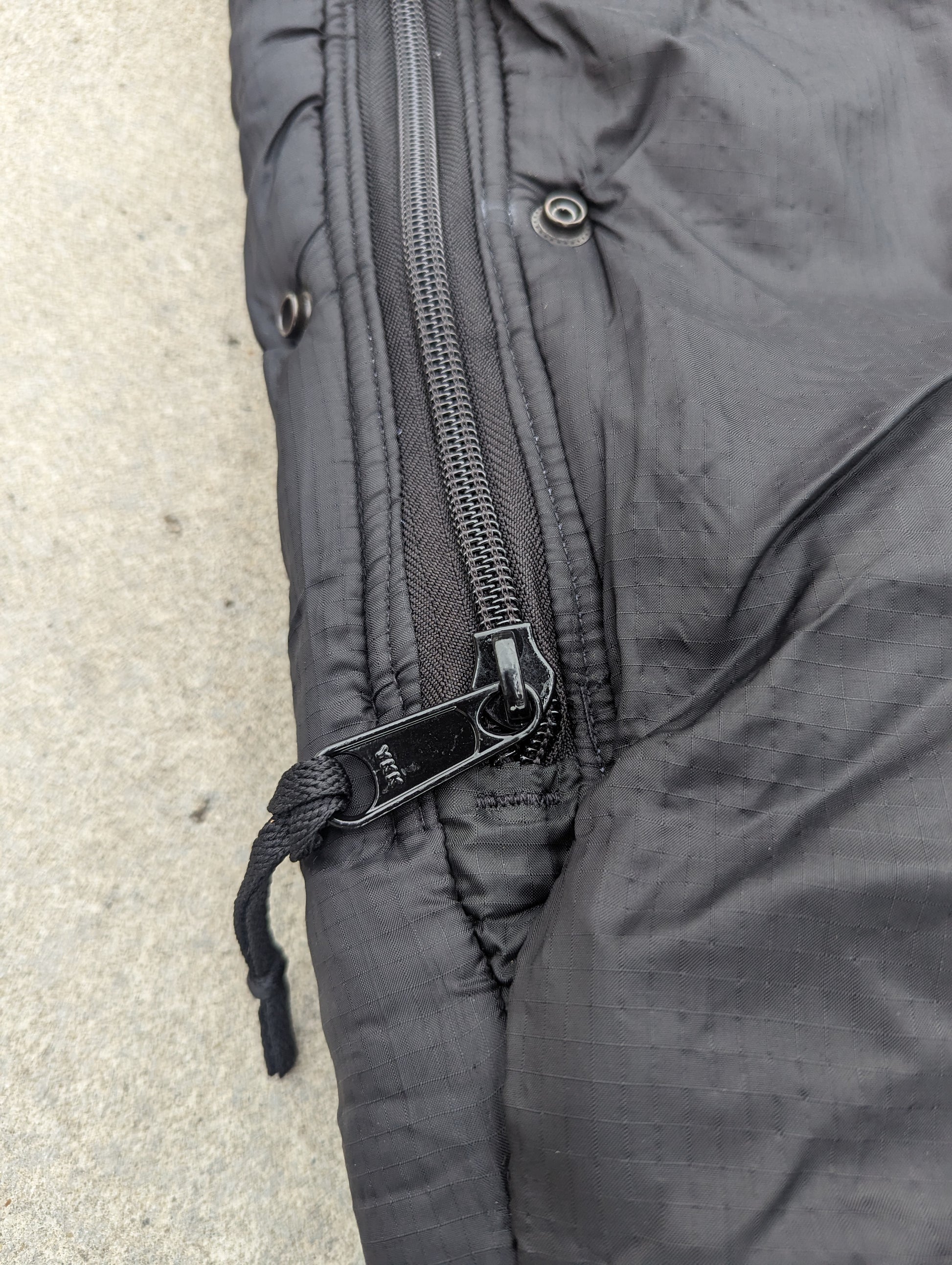A close-up detail shot of the heavy-duty zipper on a black U.S. Military Intermediate Cold Weather Sleeping Bag, showing the metal zipper pull, paracord pull, and metal snap buttons for the storm flap closure, resting on a concrete surface.