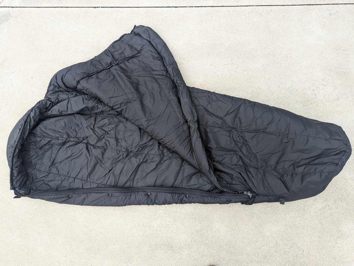 A full-length U.S. Military Intermediate Cold Weather Sleeping Bag in black, partially unzipped and laid flat on a concrete surface, displaying its quilted mummy shape and internal collar.