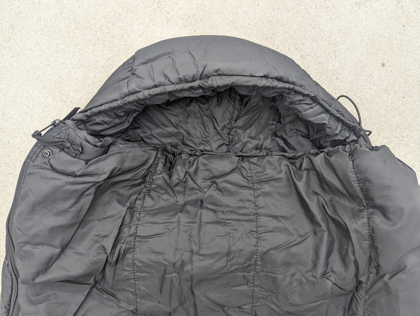 A close-up, top-down view of a U.S. Military Intermediate Cold Weather Sleeping Bag in black, showing the cinched mummy hood, quilted interior, and collar.