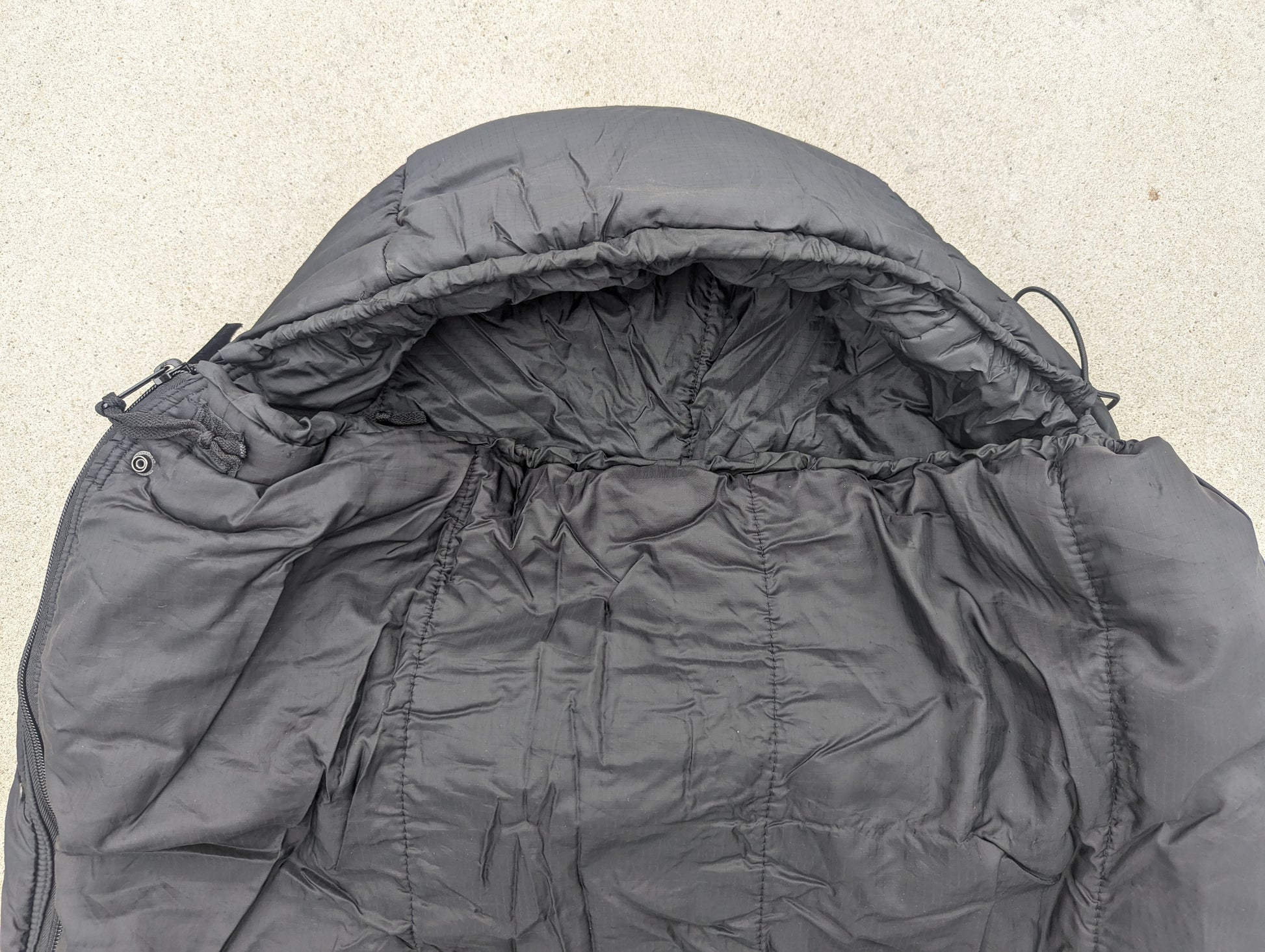 A close-up, top-down view of a U.S. Military Intermediate Cold Weather Sleeping Bag in black, showing the cinched mummy hood, quilted interior, and collar.