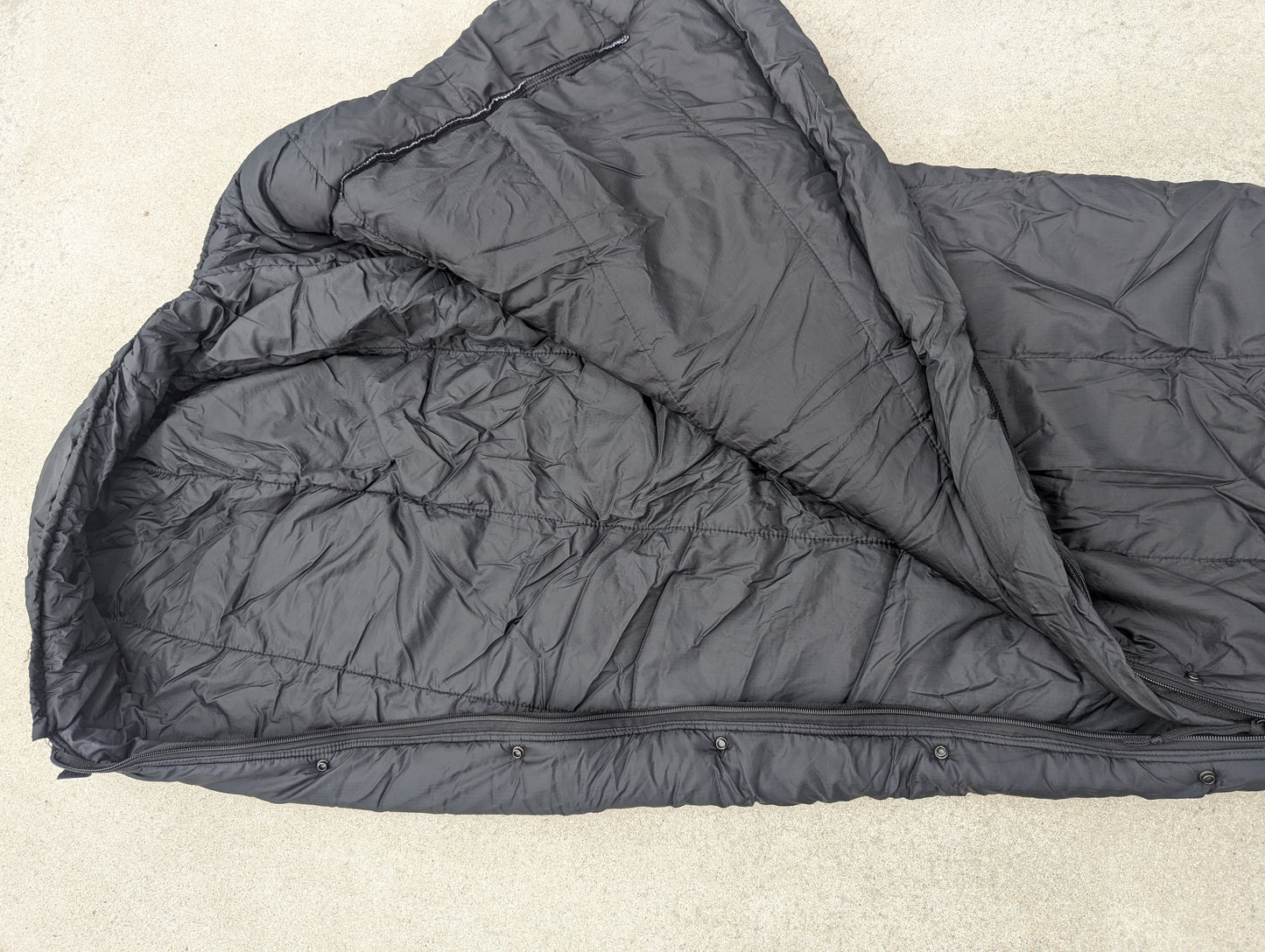 A full-length U.S. Military Intermediate Cold Weather Sleeping Bag in black, laid flat and unzipped on a concrete surface, displaying its quilted mummy shape and internal zipper flap with snaps.