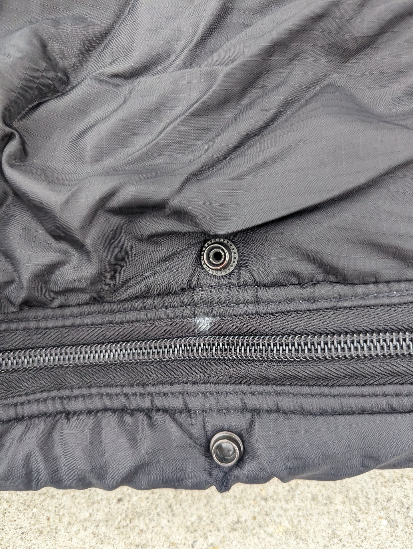 An extreme close-up detail shot of the closure on a black U.S. Military Intermediate Cold Weather Sleeping Bag, highlighting the heavy-duty zipper and the metal snap buttons for the storm flap.