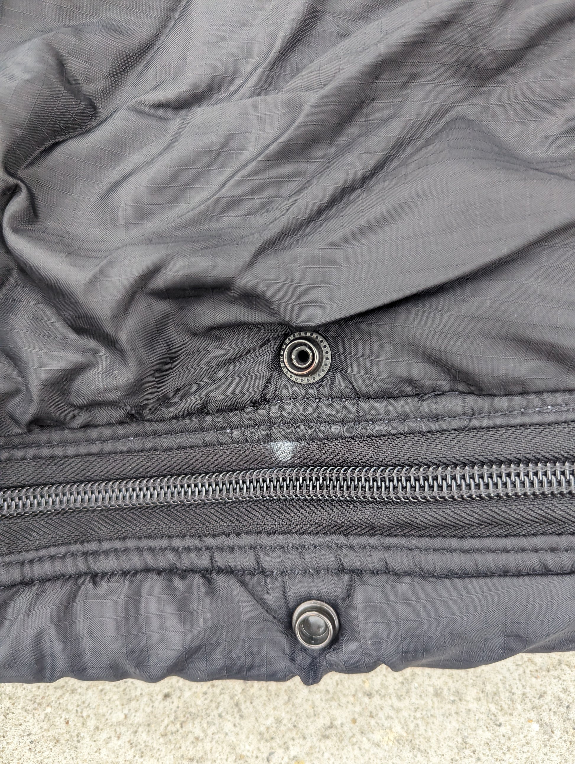 An extreme close-up detail shot of the closure on a black U.S. Military Intermediate Cold Weather Sleeping Bag, highlighting the heavy-duty zipper and the metal snap buttons for the storm flap.