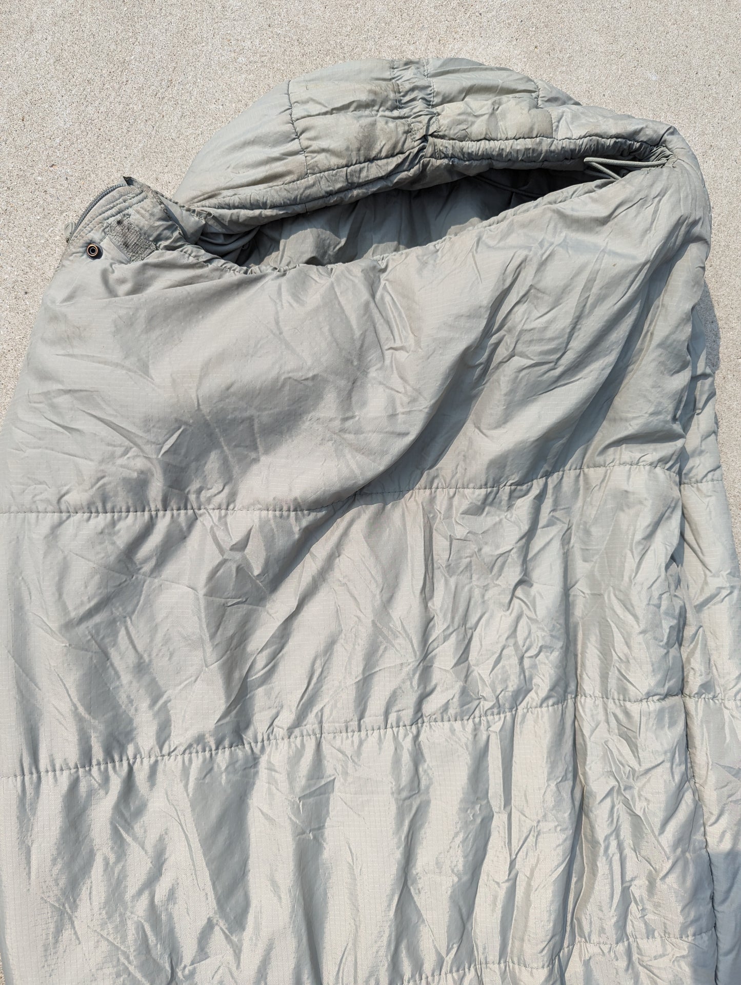 A close-up view of the hood and upper body of a U.S. Military Intermediate Cold Weather Sleeping Bag in foliage green/gray, displaying the quilted fabric texture, horizontal stitching, and cinched mummy hood.