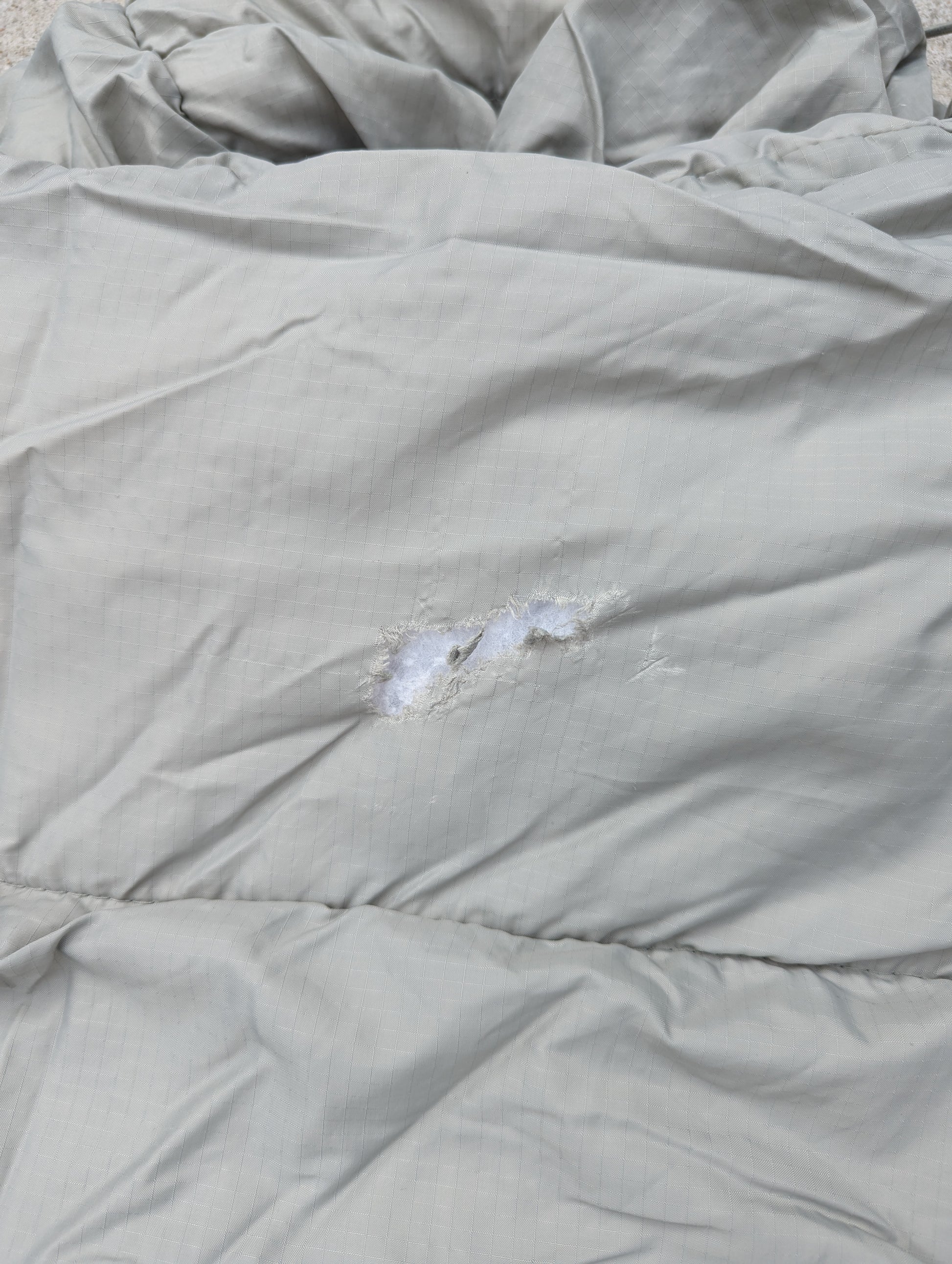 A close-up photograph of the quilted foliage green fabric of a military sleeping bag, highlighting a tear with white insulation visible, documenting damage.