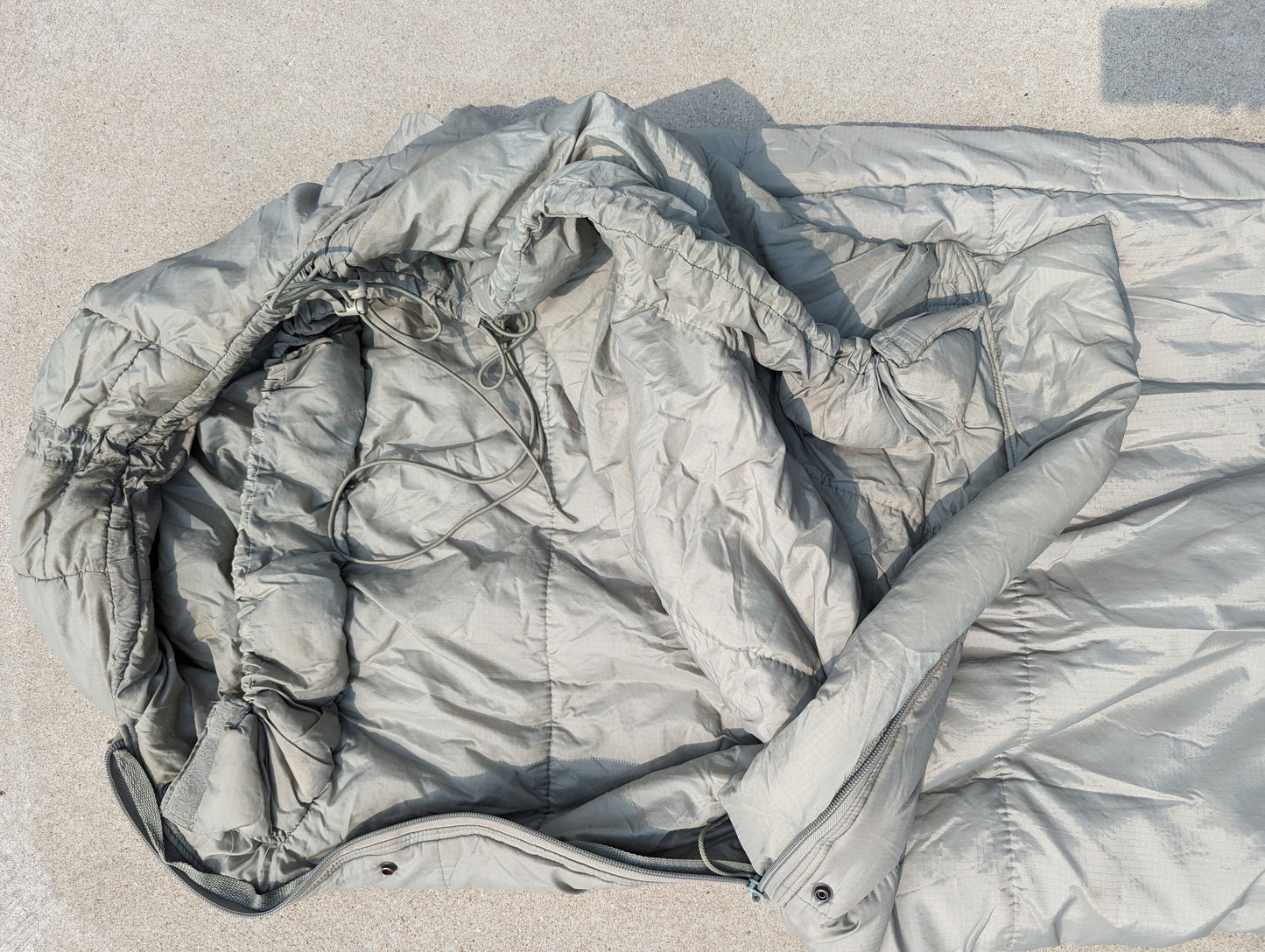 A close-up view of the hood and upper body of a U.S. Military Intermediate Cold Weather Sleeping Bag in foliage green/gray, displaying the quilted fabric, cinched drawstrings, and zipper closure.