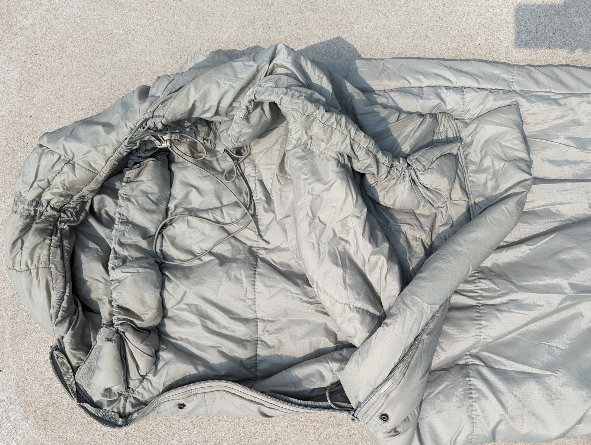 A close-up view of the hood and upper body of a U.S. Military Intermediate Cold Weather Sleeping Bag in foliage green/gray, displaying the quilted fabric, cinched drawstrings, and zipper closure.
