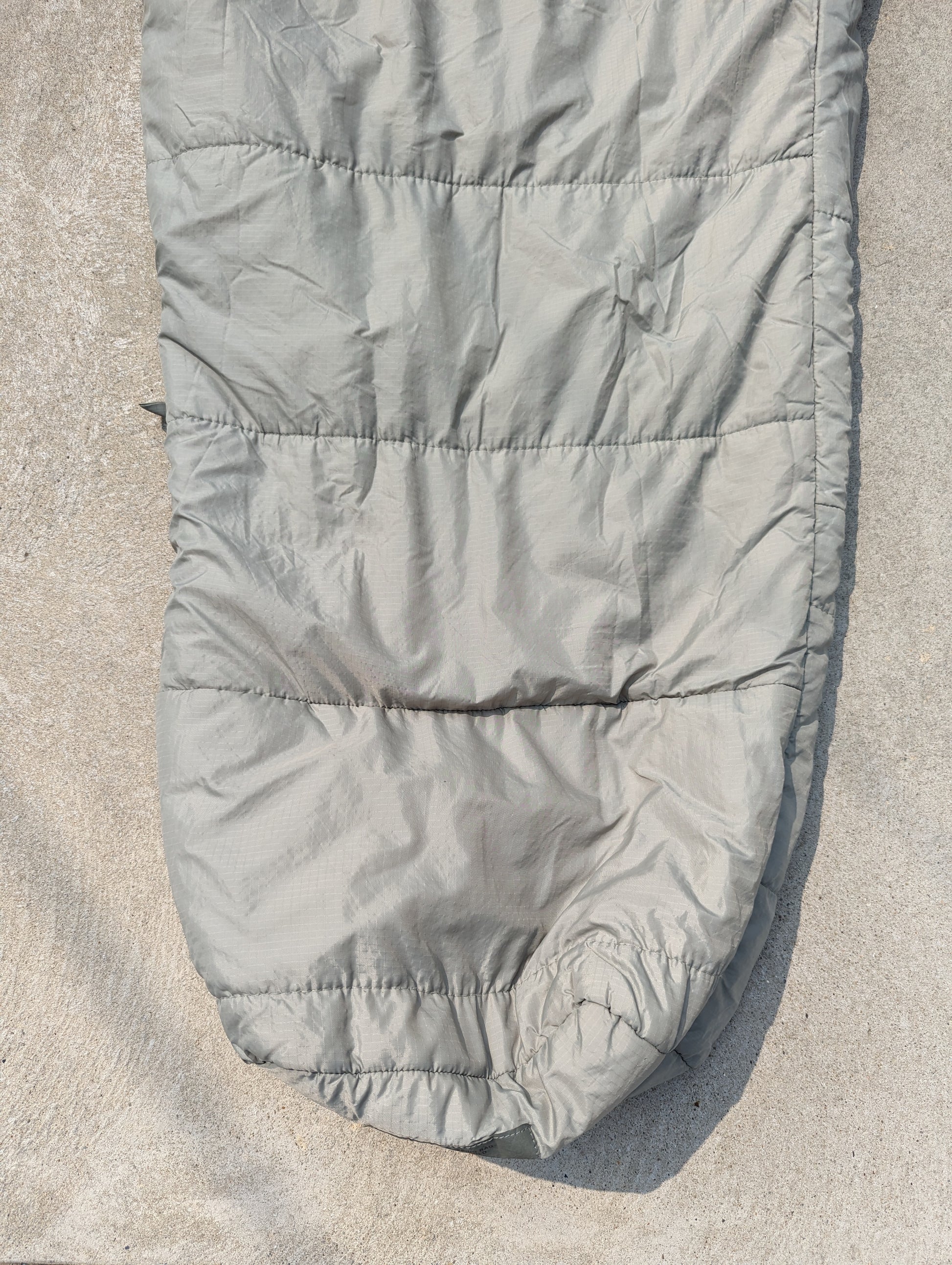 A full-length U.S. Military Surplus Intermediate Cold Weather Sleeping Bag in foliage green/gray, laid flat on a textured surface, displaying its horizontal quilted pattern and mummy shape.