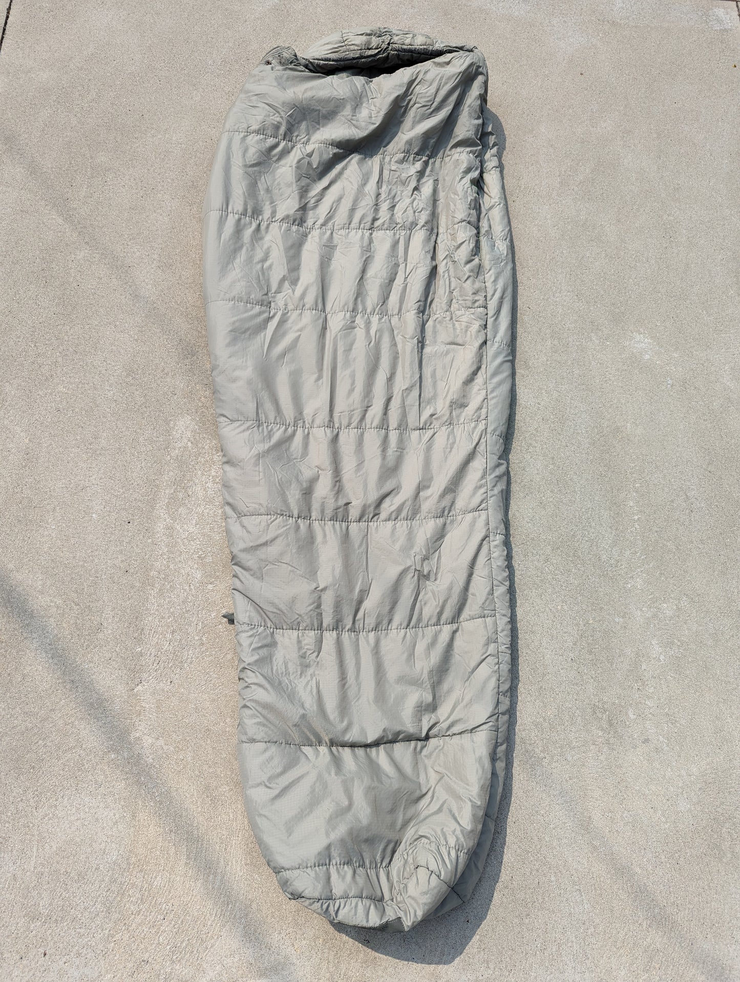 A full-length U.S. Military Surplus Intermediate Cold Weather Sleeping Bag in foliage green/gray, laid flat on a concrete surface, displaying its horizontal quilted pattern and mummy shape.