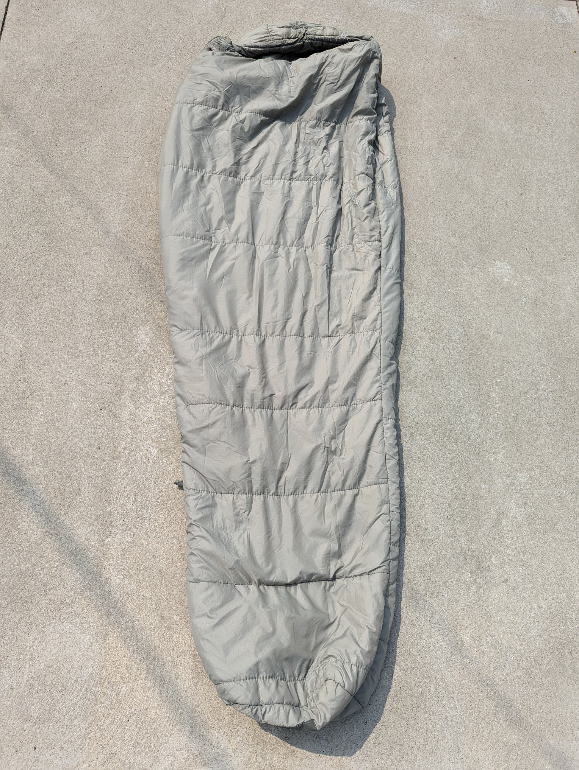 A full-length U.S. Military Surplus Intermediate Cold Weather Sleeping Bag in foliage green/gray, laid flat on a concrete surface, displaying its horizontal quilted pattern and mummy shape.