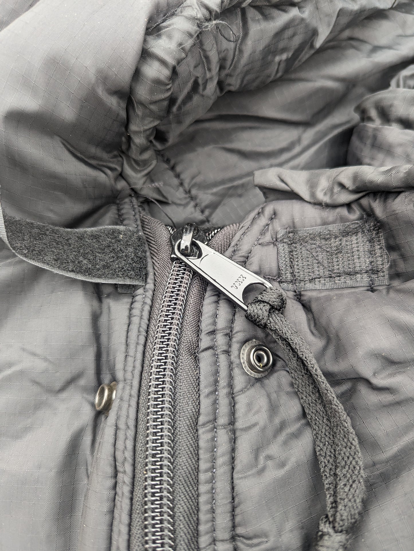 An extreme close-up of the dark gray quilted fabric on a U.S. Military Intermediate Cold Weather Sleeping Bag, highlighting the metal zipper pull, hook-and-loop tab, and hood drawstring.