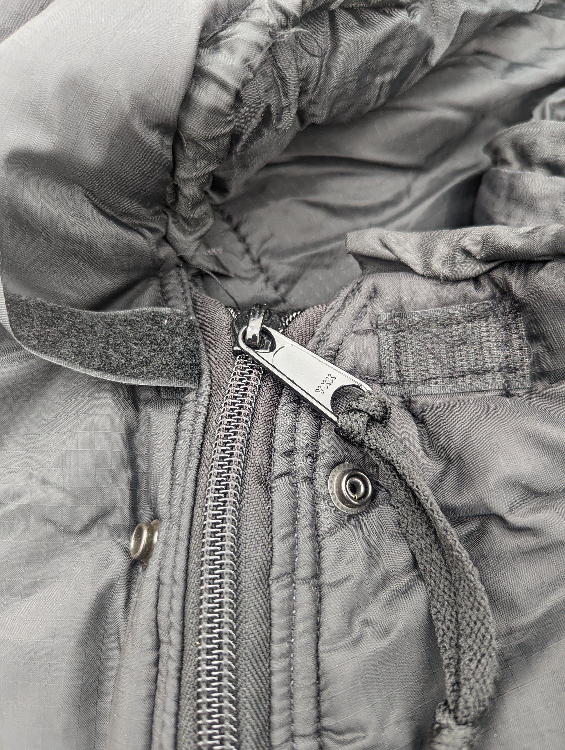 An extreme close-up of the dark gray quilted fabric on a U.S. Military Intermediate Cold Weather Sleeping Bag, highlighting the metal zipper pull, hook-and-loop tab, and hood drawstring.