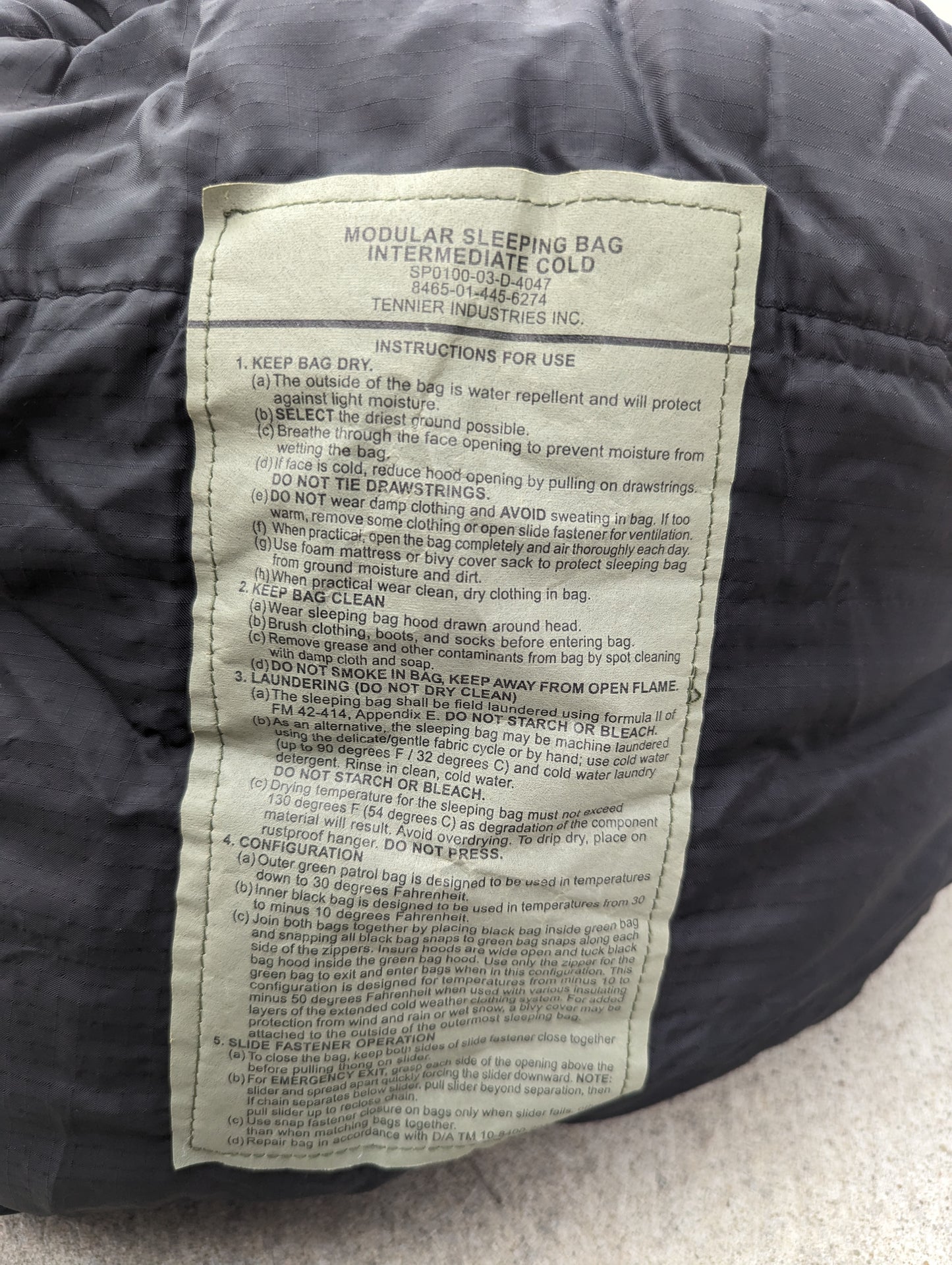 A close-up photograph of the official U.S. Military issue tag sewn into the Intermediate Cold Weather Sleeping Bag, clearly displaying the product name, instructions for use, care, cleaning, and storage, and manufacturer details.