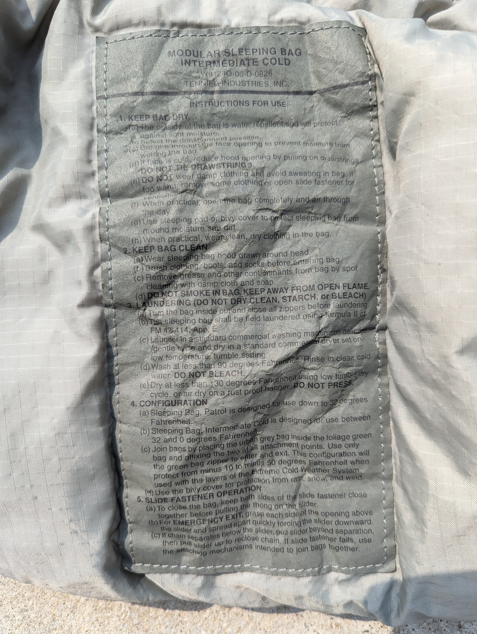 A close-up photograph of the large official U.S. Military issue tag sewn into the Intermediate Cold Weather Sleeping Bag, clearly displaying the product name 'MODULAR SLEEPING BAG INTERMEDIATE COLD' and sections for instructions for use, care, and maintenance.