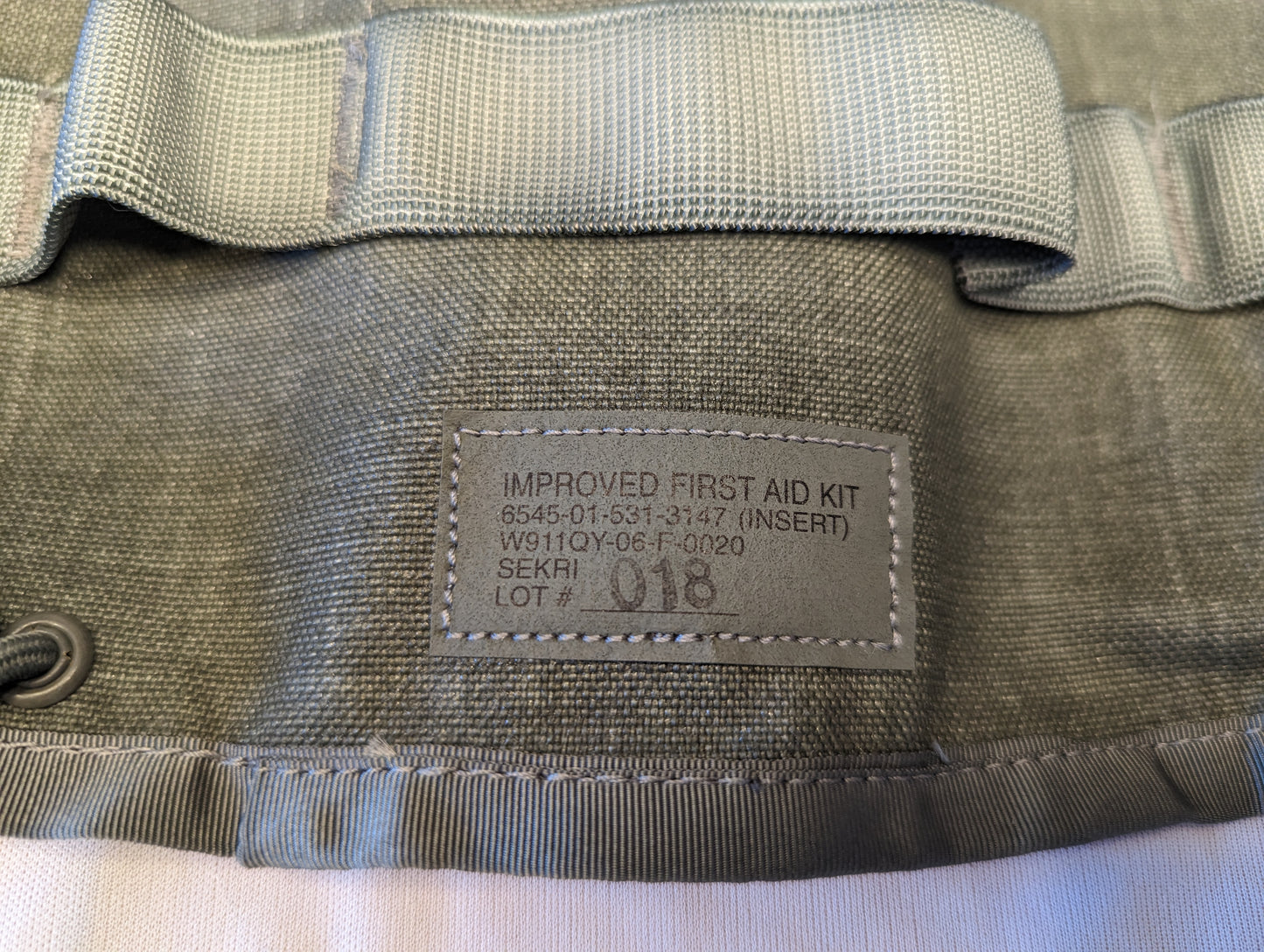 A close-up photograph of the official U.S. Military issue tag sewn into the olive green IFAK insert, displaying 'IMPROVED FIRST AID KIT (INSERT)', NSN 8465-01-531-3147, and manufacturer SEKRI.