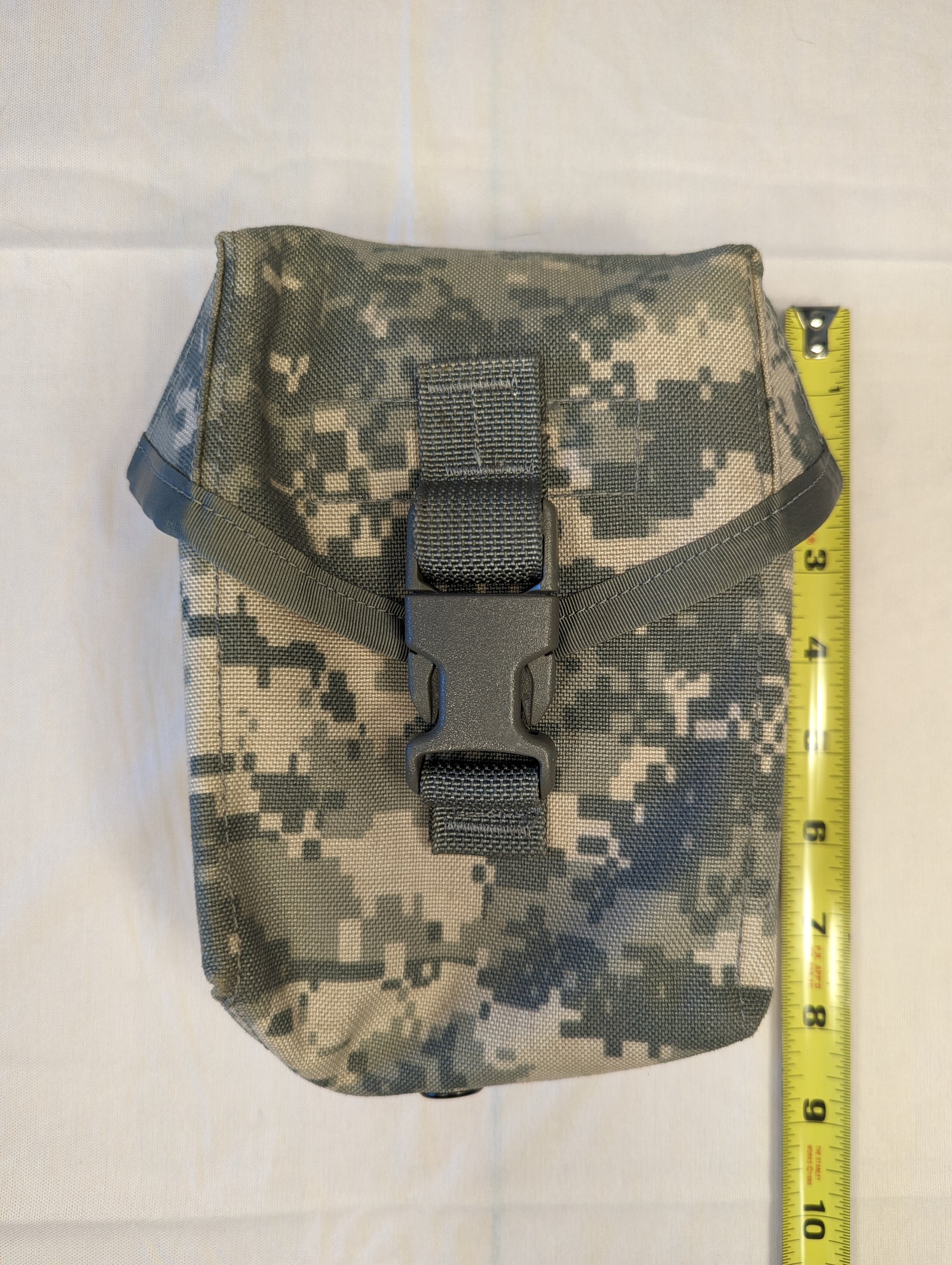 A single U.S. Military Surplus Improved First Aid Kit (IFAK) Pouch in ACU digital camouflage, standing upright next to a tape measure to demonstrate its height, secured with a black quick-release buckle.