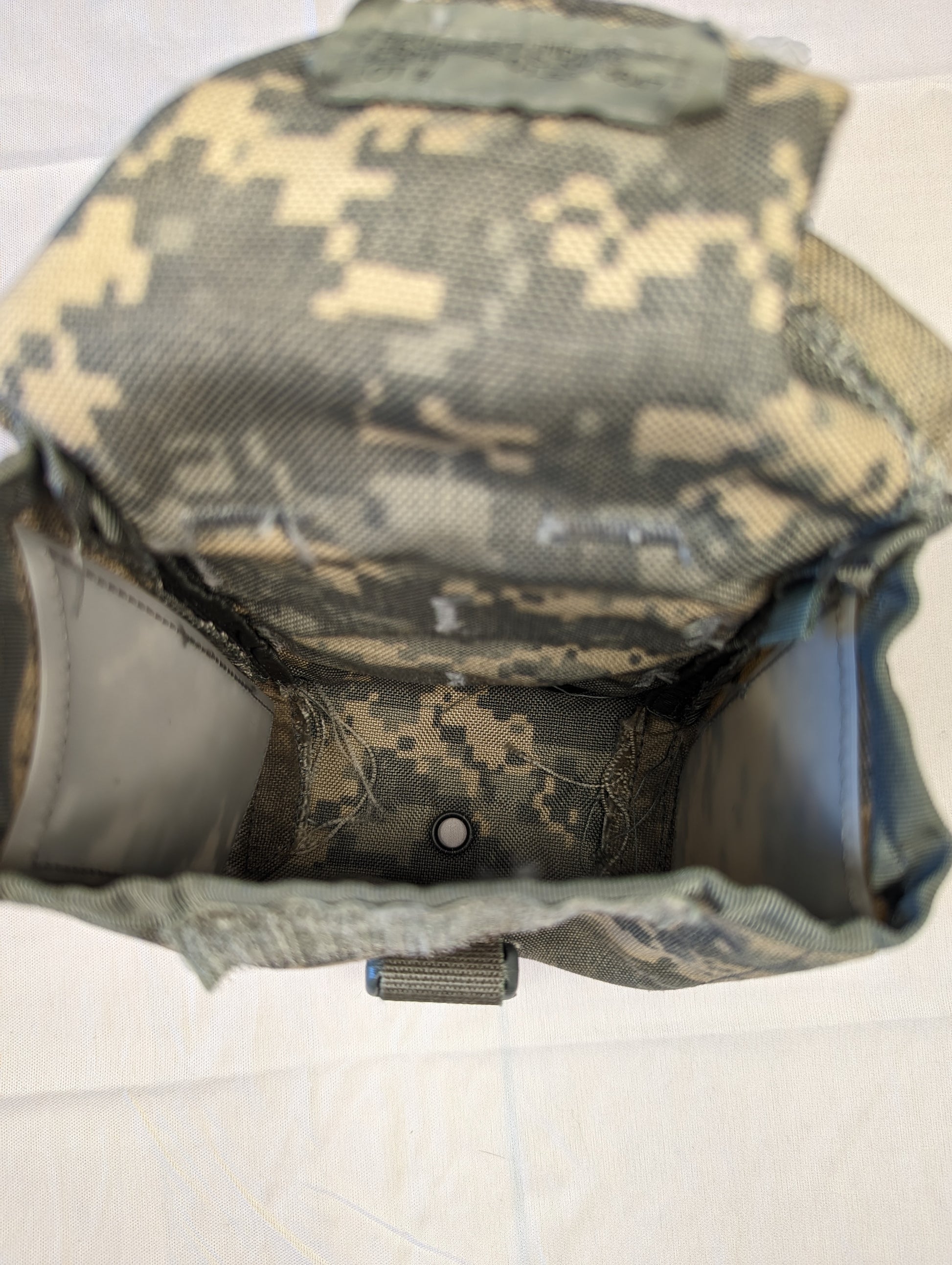 A high-angle view looking deep inside the empty UCP/ACU Individual First Aid Kit (IFAK) pouch, highlighting the dark, soiled interior and the metal drainage grommet on the floor.