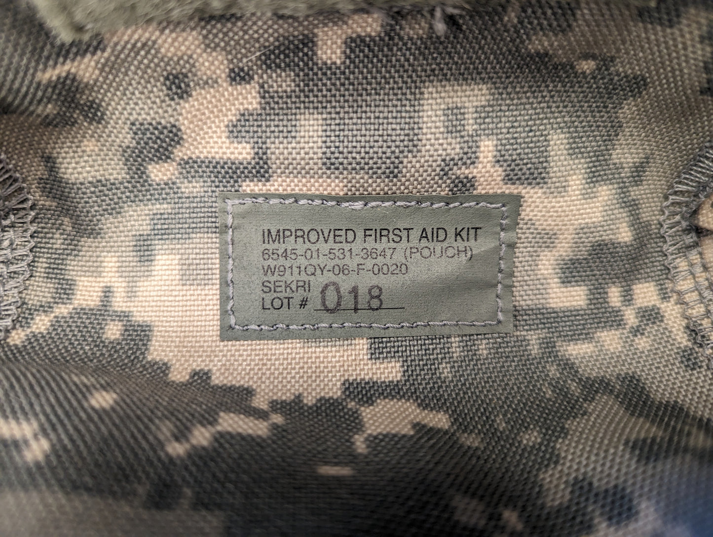 A close-up photograph of the official U.S. Military issue tag sewn into the UCP/ACU Individual First Aid Kit pouch, displaying 'IMPROVED FIRST AID KIT (POUCH)', NSN 8465-01-531-3647, and 'LOT # 018' markings.