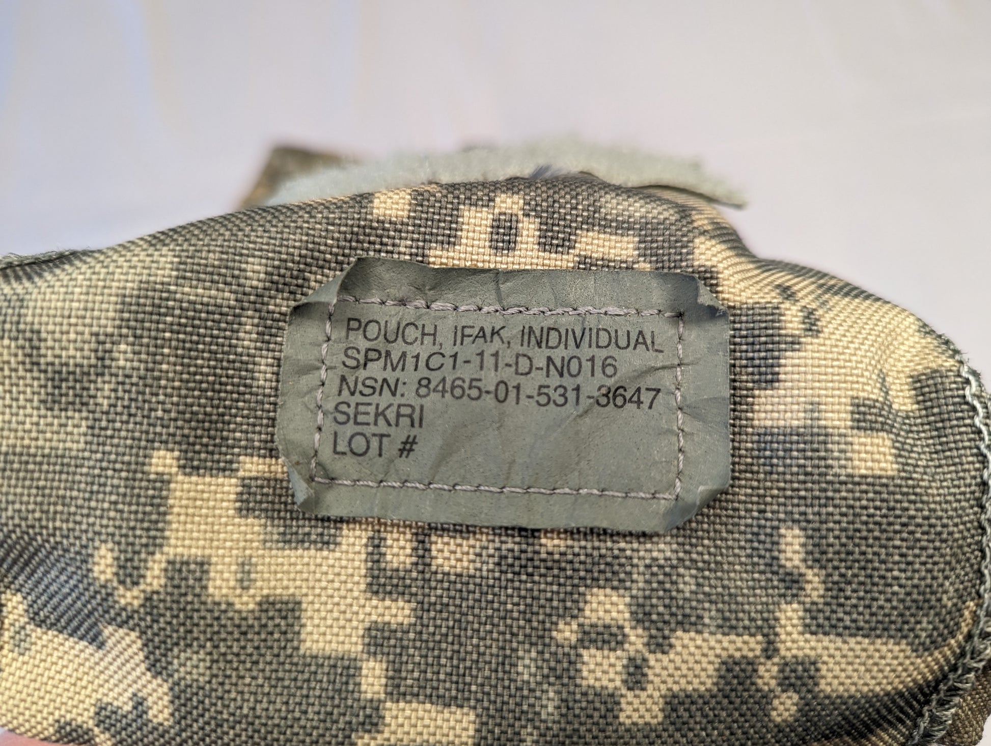 A close-up photograph of the official U.S. Military issue tag sewn into the UCP/ACU IFAK pouch, displaying the product name 'POUCH, IFAK, INDIVIDUAL', NSN 8465-01-531-3647, and manufacturer SEKRI.