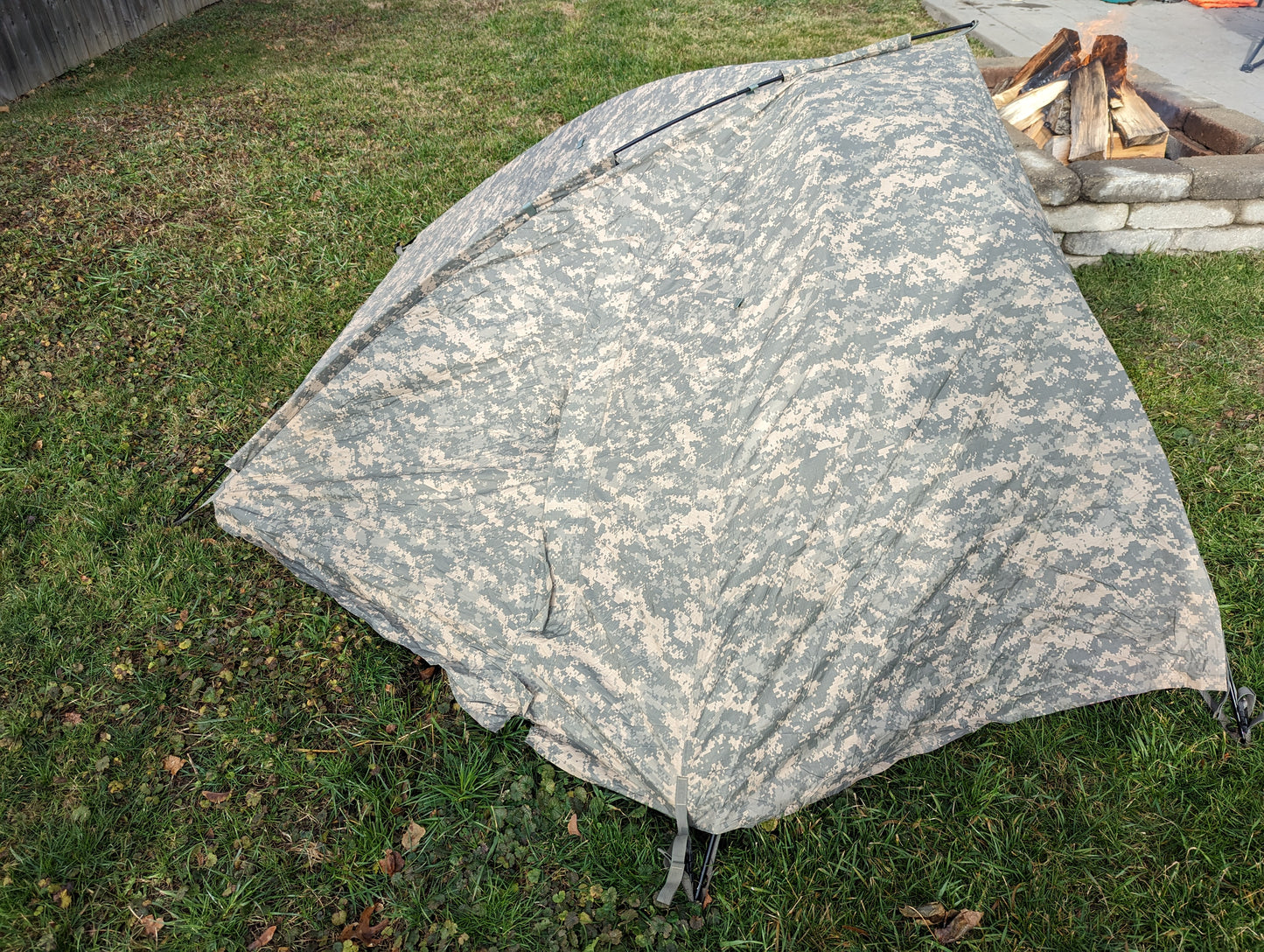 A U.S. Military Improved Combat Shelter (ICS) tent pitched on a grassy lawn, featuring an ACU digital camouflage rainfly, positioned near an active stone fire pit.