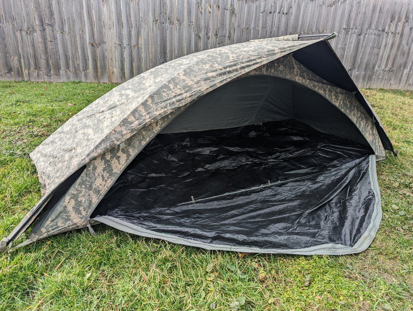 A U.S. Military Improved Combat Shelter (ICS) tent pitched outdoors on a grassy lawn, featuring an ACU digital camouflage fly with the entrance open to reveal the black waterproof floor.