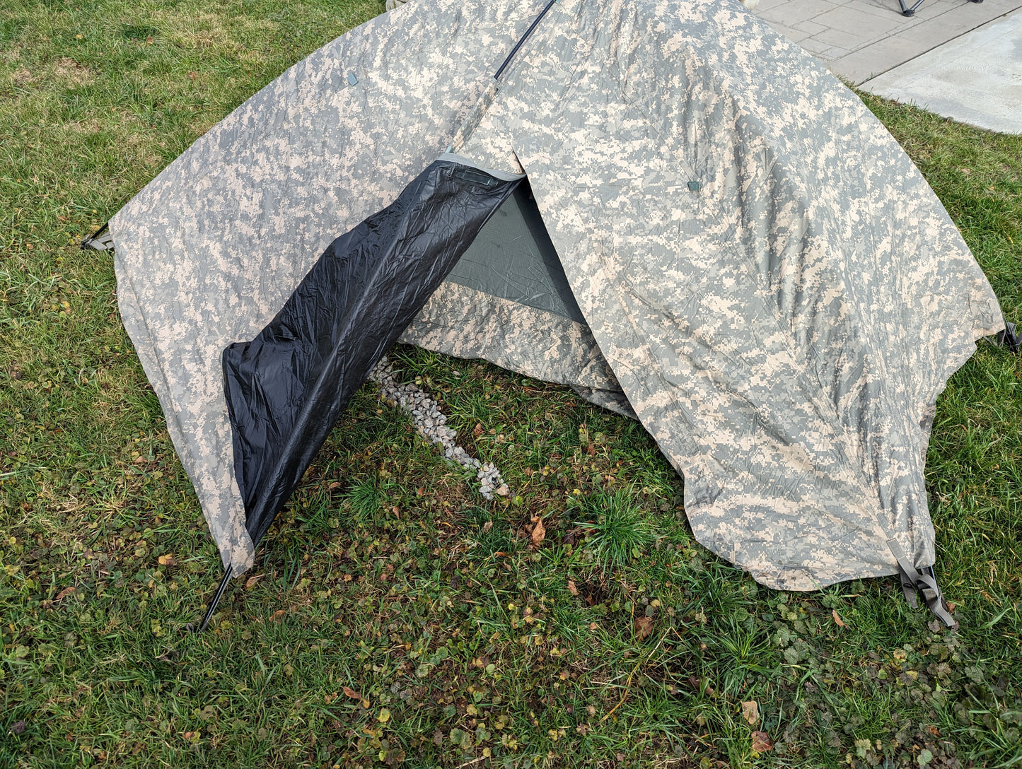 A U.S. Military Improved Combat Shelter (ICS) tent pitched outdoors on grass, featuring an ACU digital camouflage rainfly with the front flap rolled back to expose the inner mesh netting.