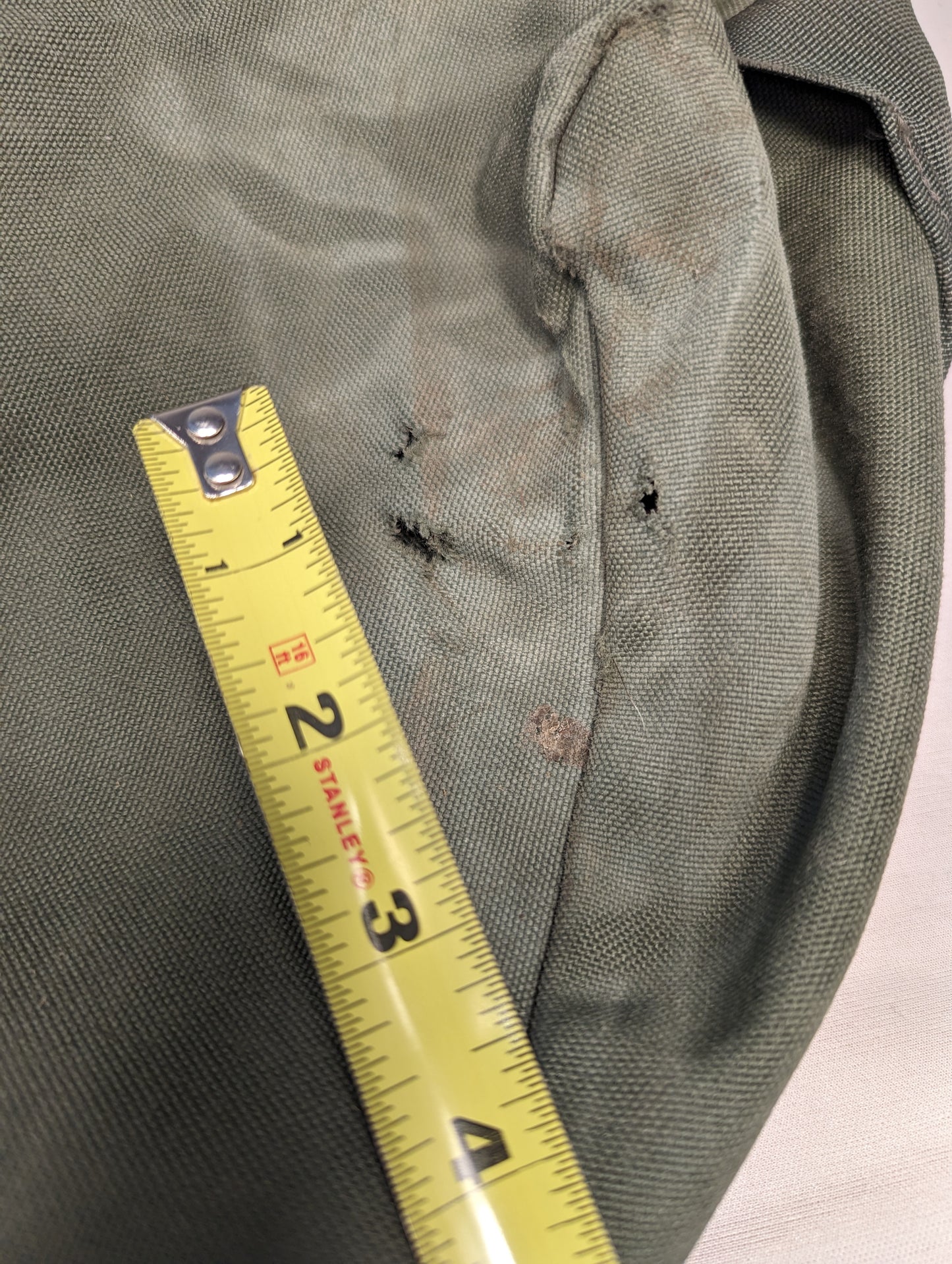 An extreme close-up detail shot of the olive green U.S. Military Improved Duffel Bag, showing multiple tears and holes in the canvas fabric, with a yellow tape measure demonstrating the extent of the damage.