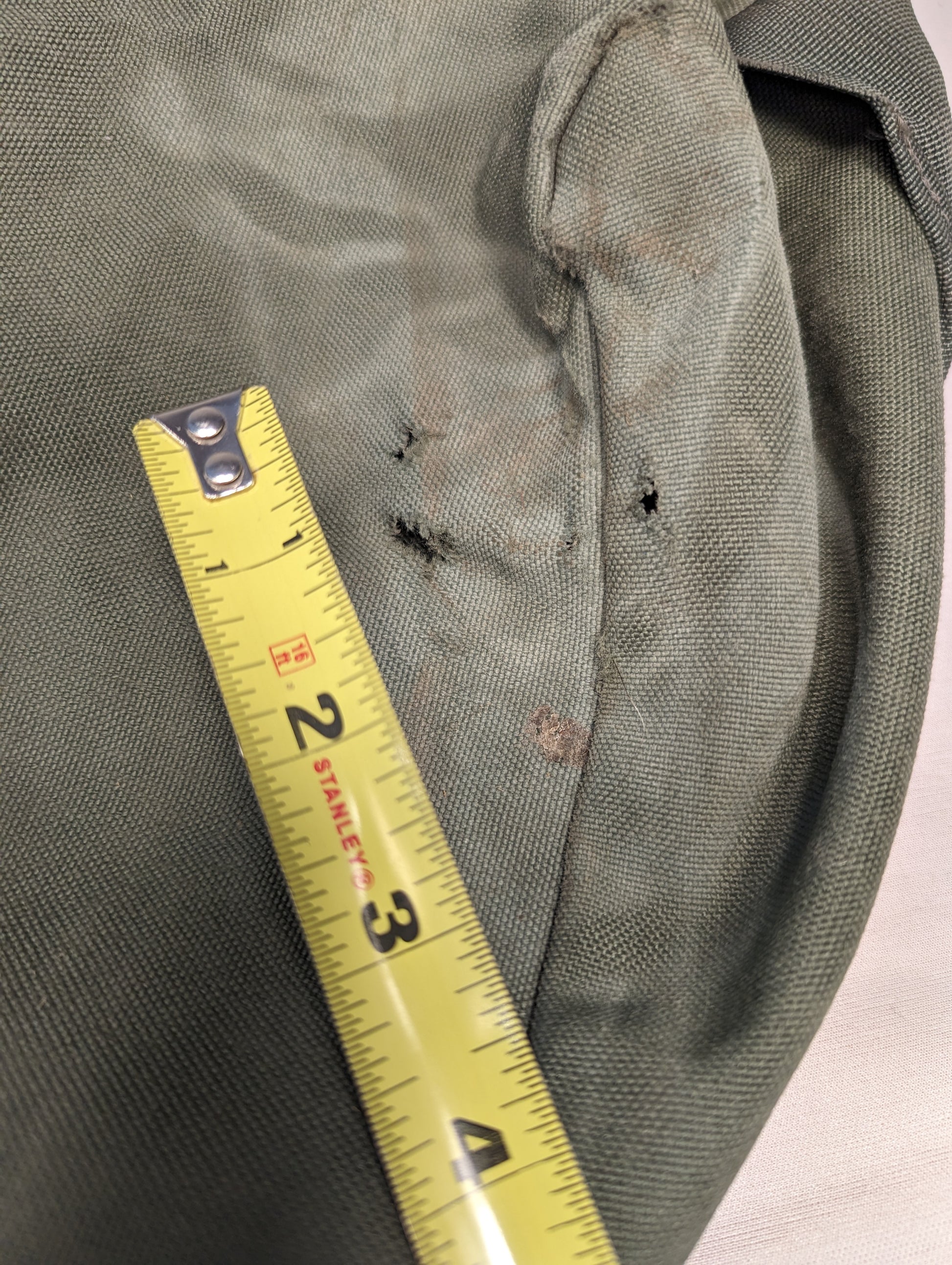 An extreme close-up detail shot of the olive green U.S. Military Improved Duffel Bag, showing multiple tears and holes in the canvas fabric, with a yellow tape measure demonstrating the extent of the damage.
