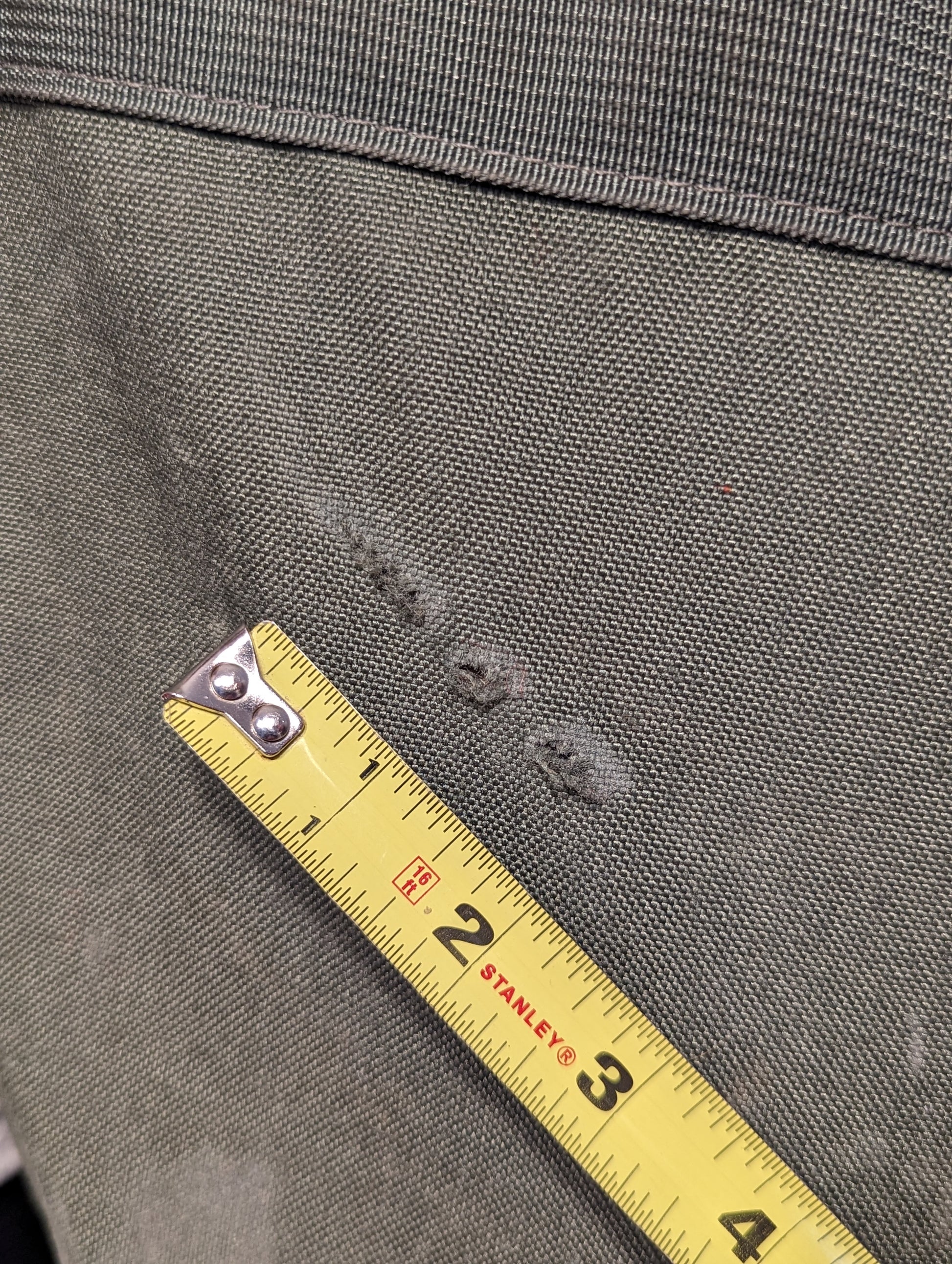 A close-up detail shot of the olive green U.S. Military Improved Duffel Bag fabric, showing three small snags or tears next to a tape measure, emphasizing the used/surplus condition.