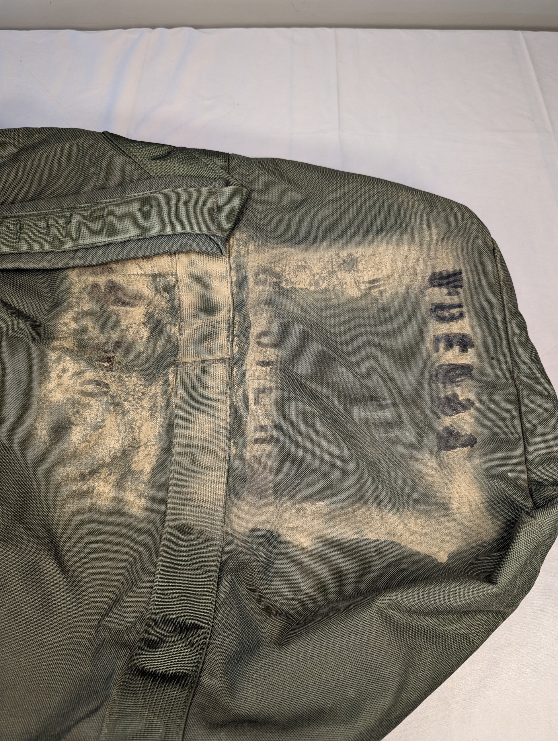 A close-up detail shot of the olive green U.S. Military Improved Duffel Bag fabric, the heavy wear, and a large, faded stenciled government marking on the side.