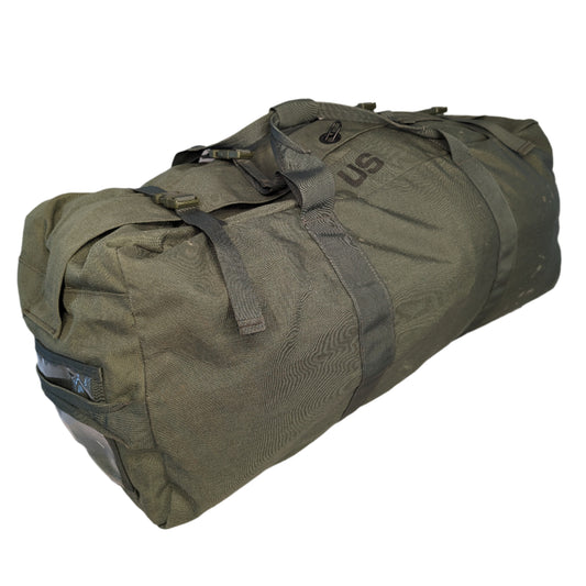 A large U.S. Military Surplus Improved Duffel Bag (IDB) in olive green, laid diagonally on a white background, featuring heavy-duty webbing straps and a prominent stenciled 'US' mark.