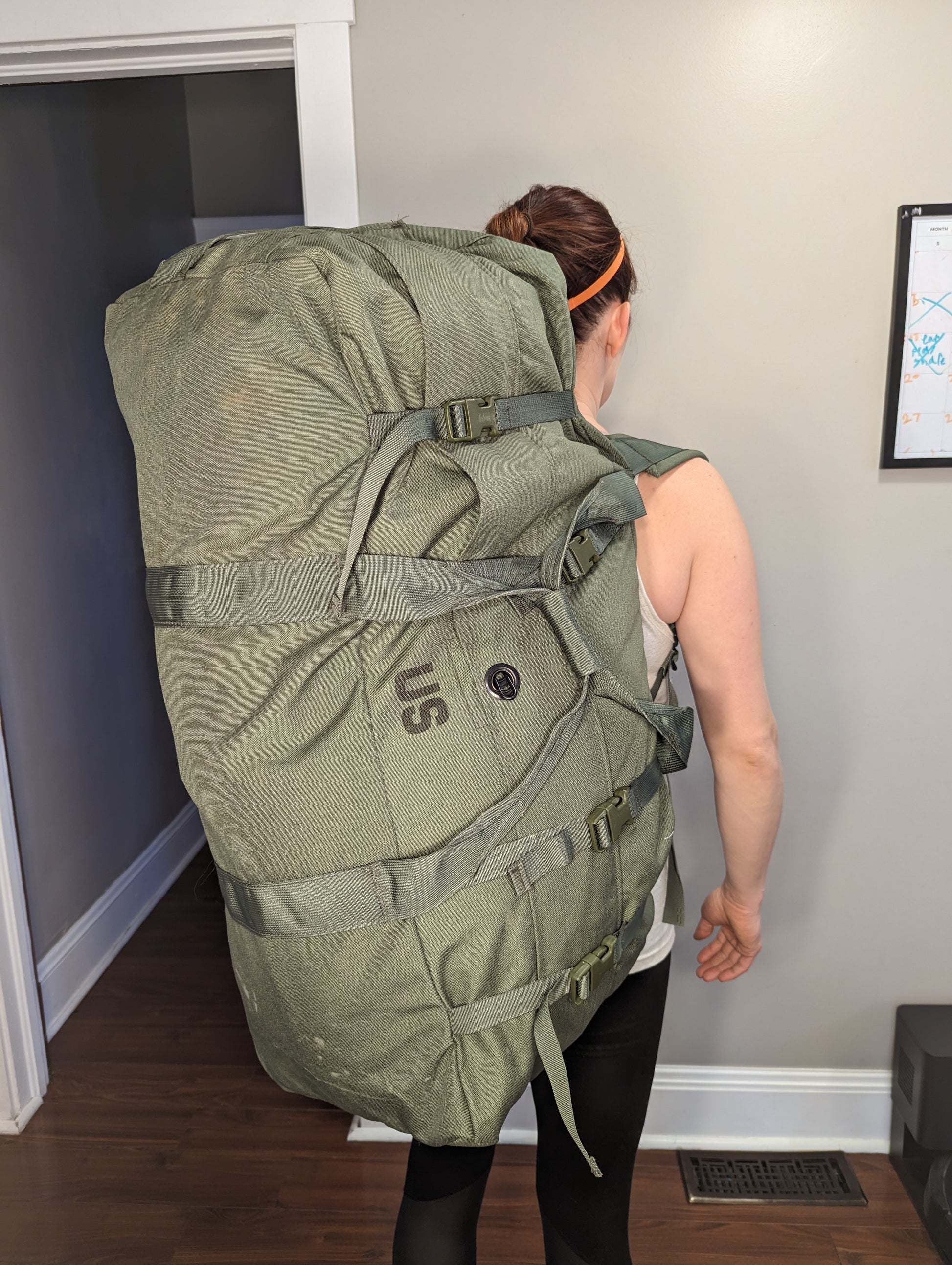 A woman carrying a very large, olive green U.S. Military Surplus Improved Duffel Bag (IDB) vertically on her back, emphasizing the bag's massive size, and the 'U.S.' government marking.