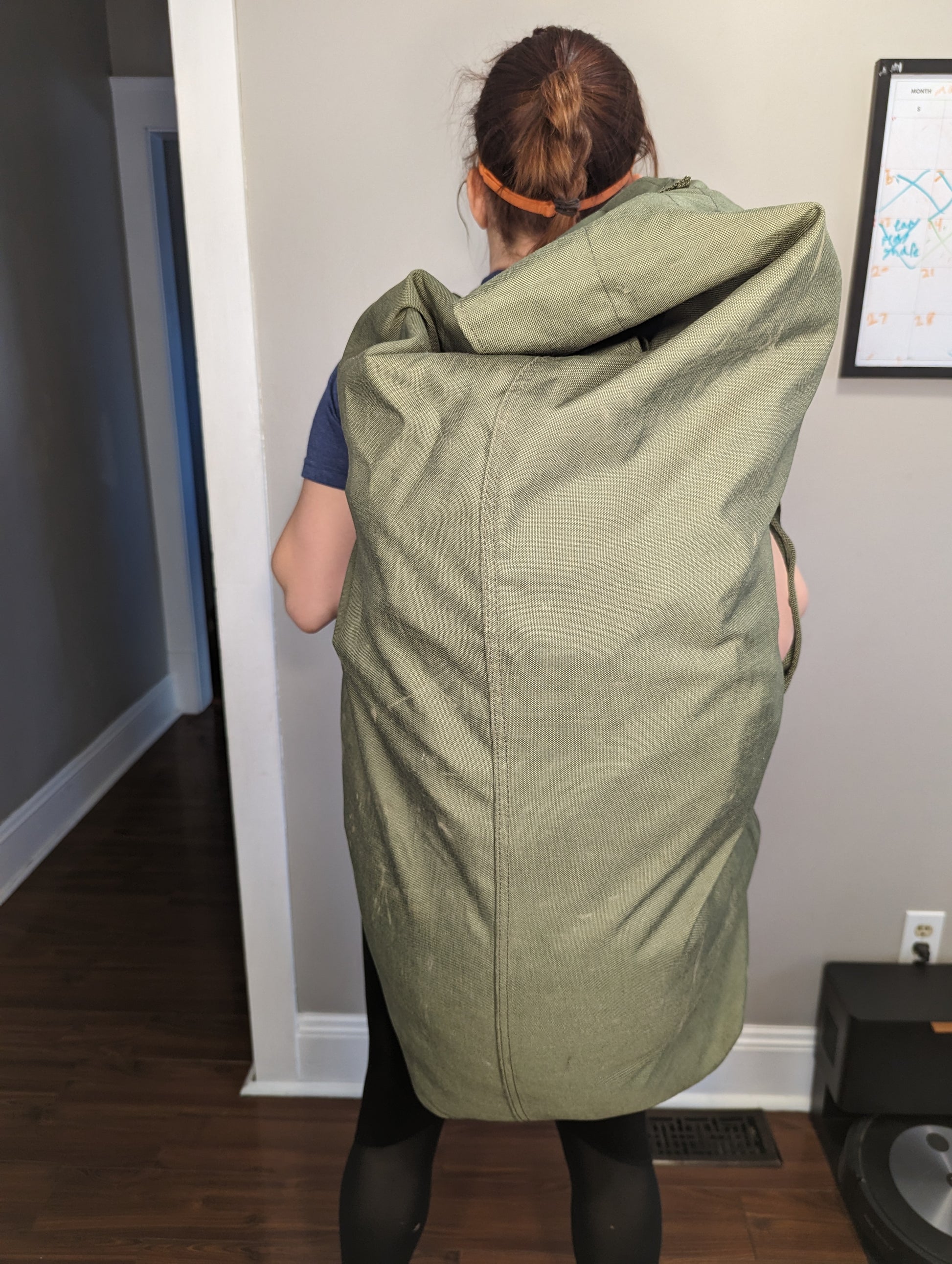 A woman carrying a very large, olive green U.S. Military Surplus Duffel Bag slung over her shoulder, showcasing the bag's immense size, wavy quilted texture, and vertical orientation against her back.