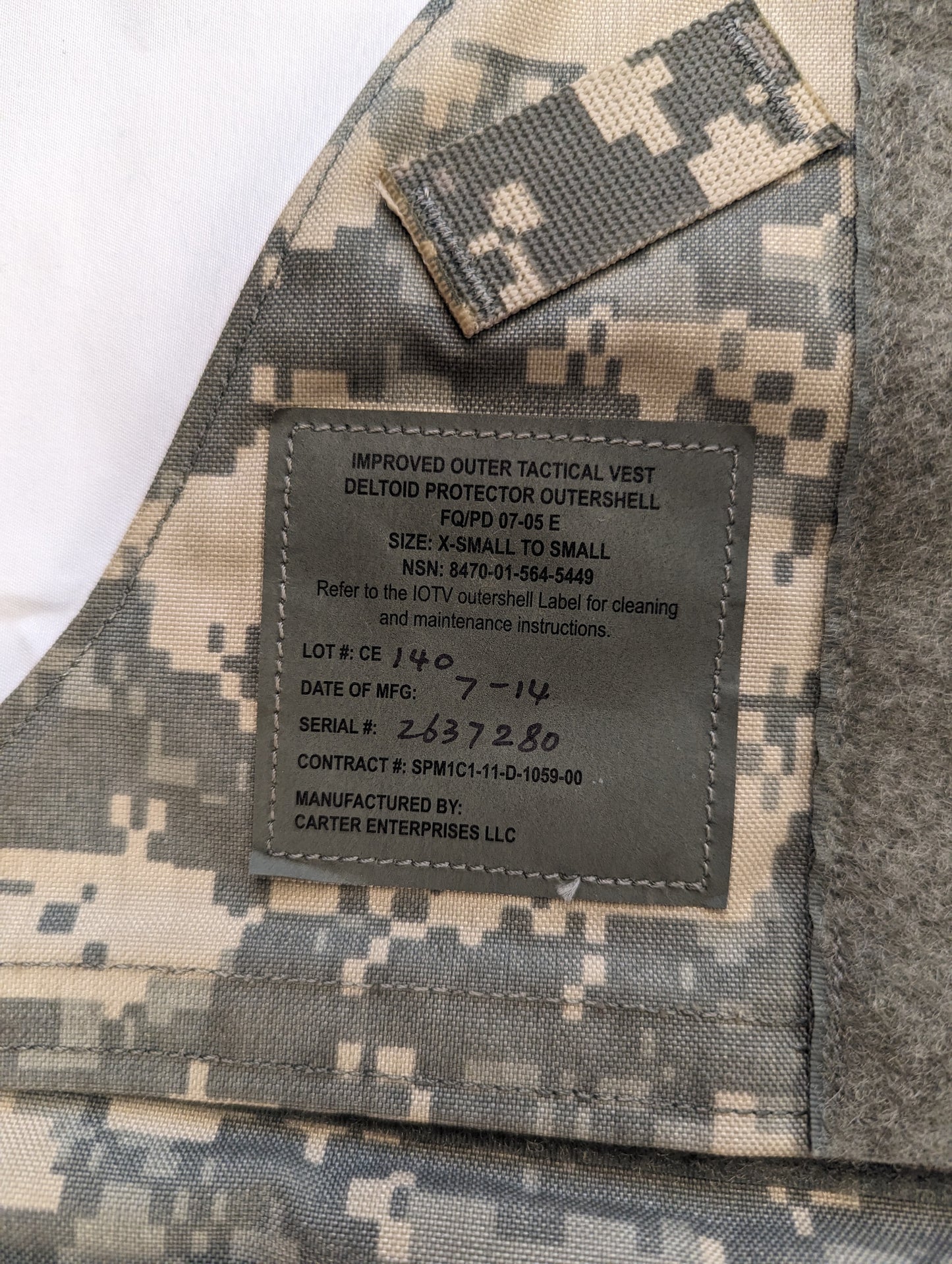 A close-up photograph of the original U.S. Military issue tag sewn into the ACU camouflage IOTV, displaying the product name, contract number, size information, and date of manufacture.