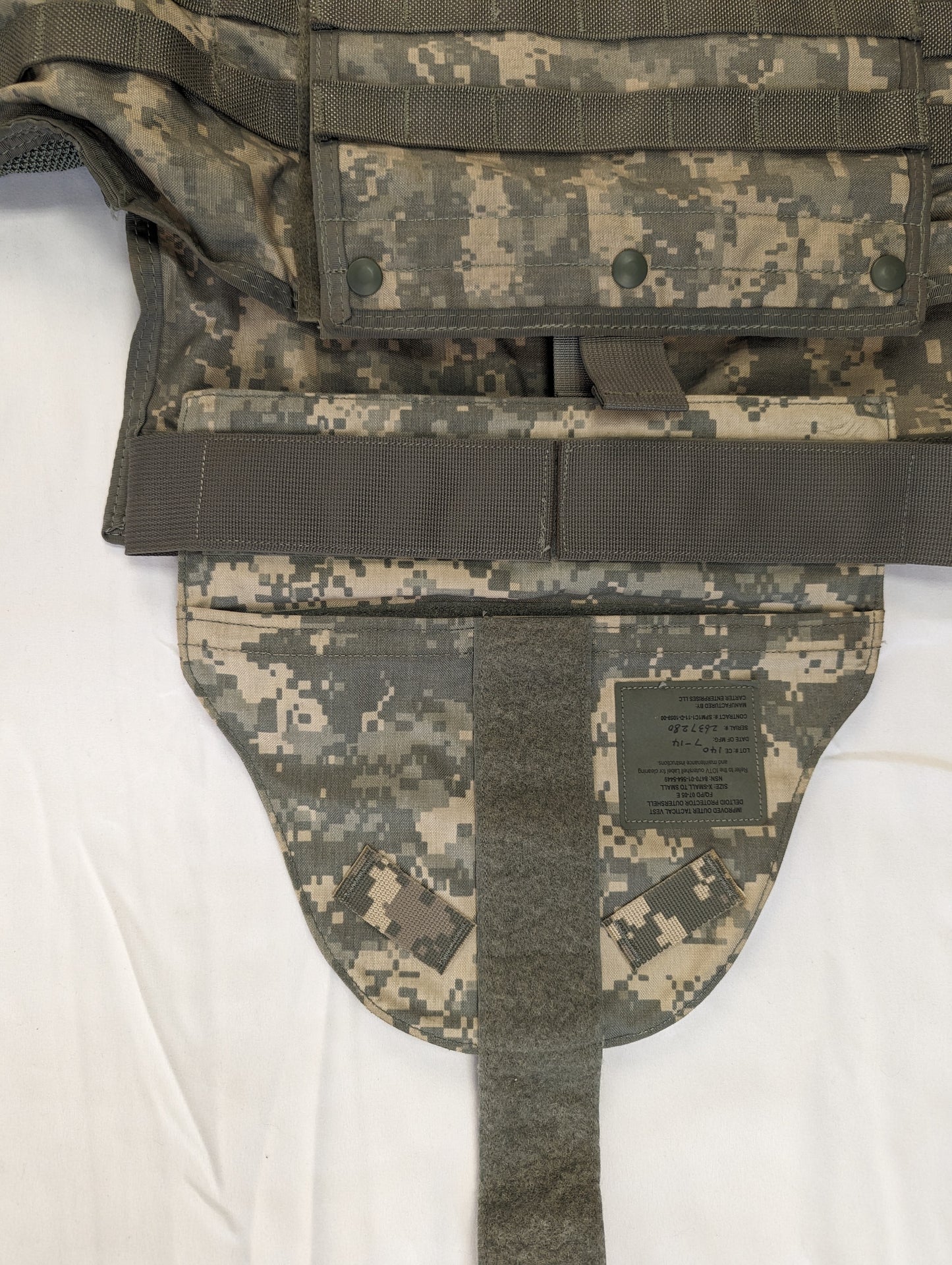 A close-up of the front lower torso of an ACU camouflage IOTV, highlighting the adjustable cummerbund strap, the hook-and-loop closure, and the 'GROIN PROTECTOR' issue tag.