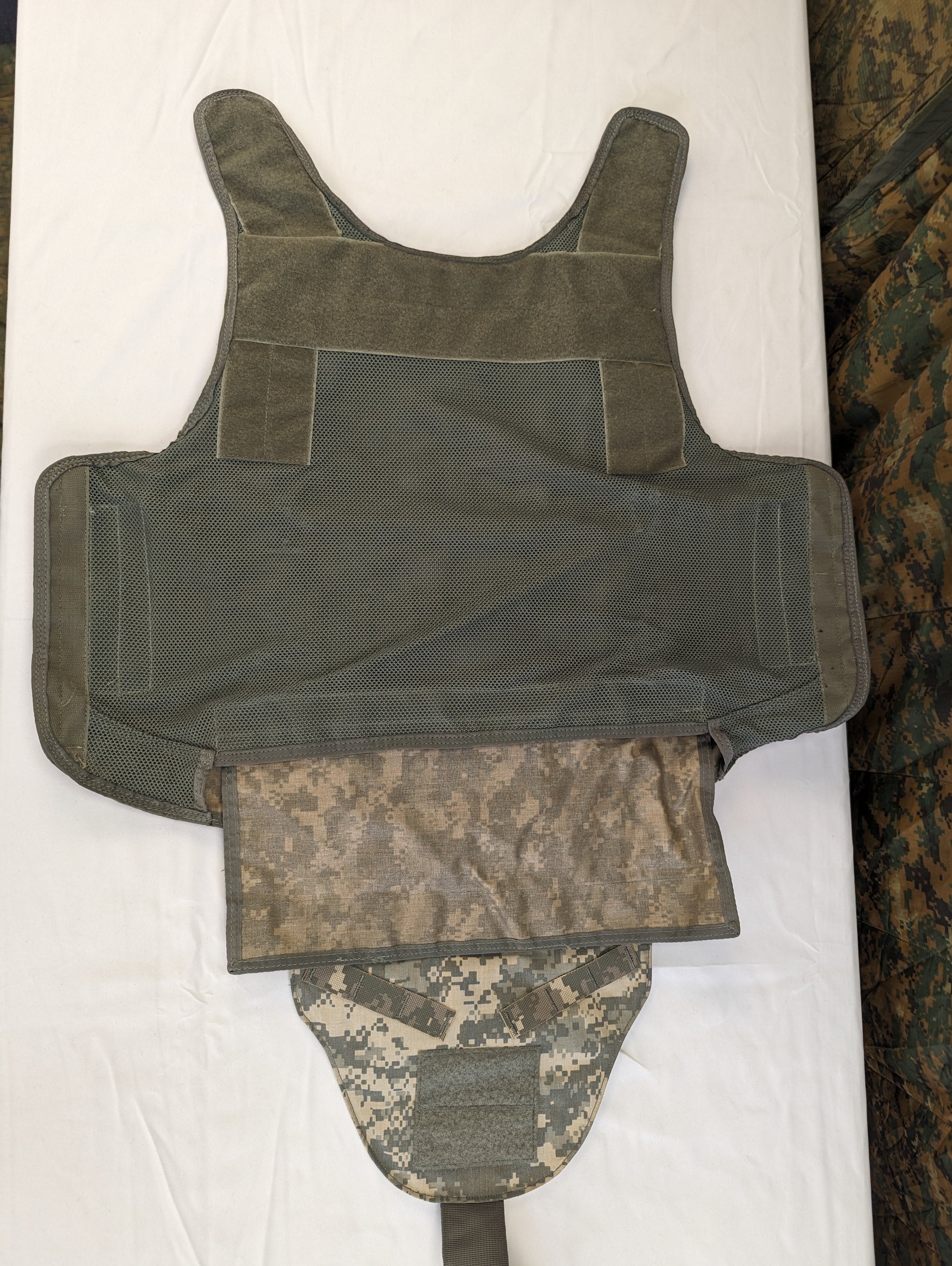 A close-up view of the internal components of the Improved Outer Tactical Vest (IOTV), showing the quilted olive drab main body and the attached ACU camouflage groin protector flap.
